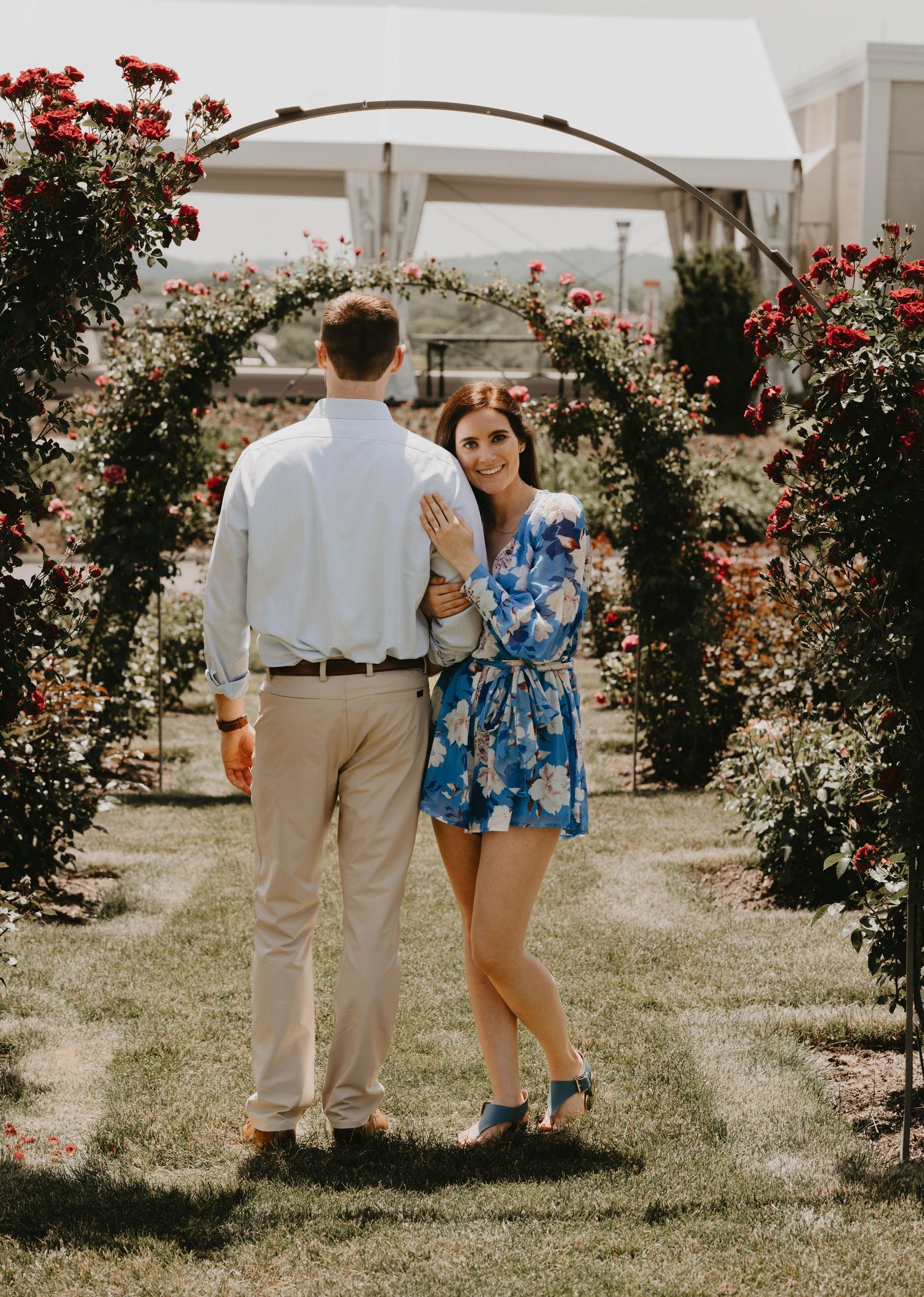 A couple poses under a floral archway in a garden setting with red roses and greenery.