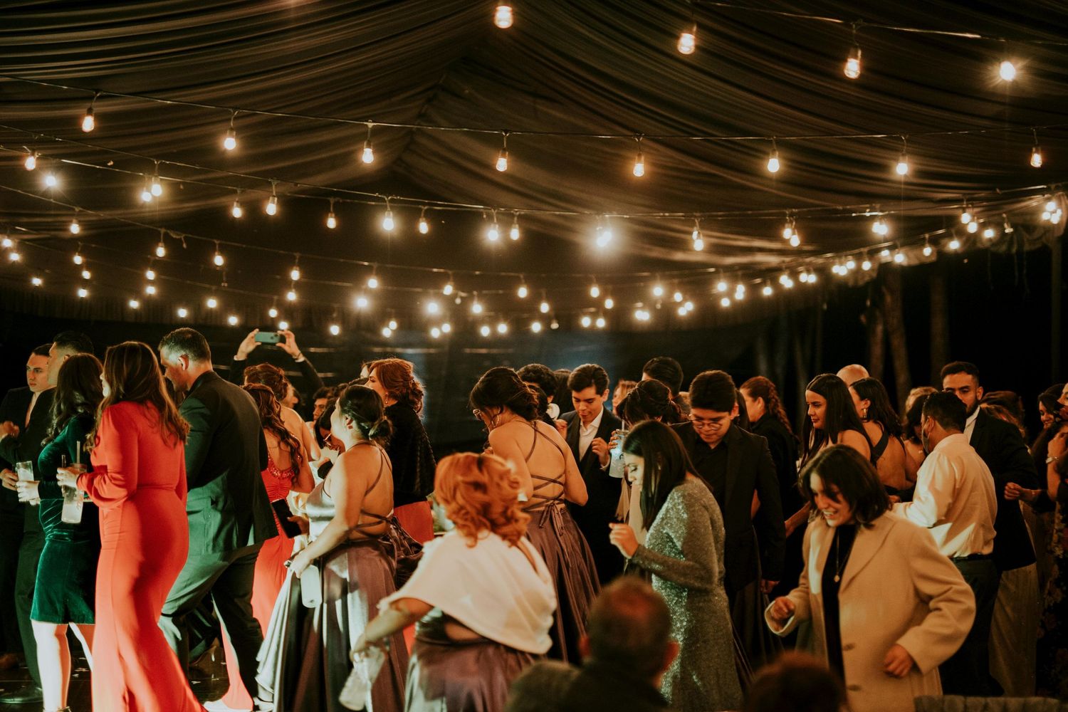 Large group of people dancing under string lights at an outdoor evening celebration.