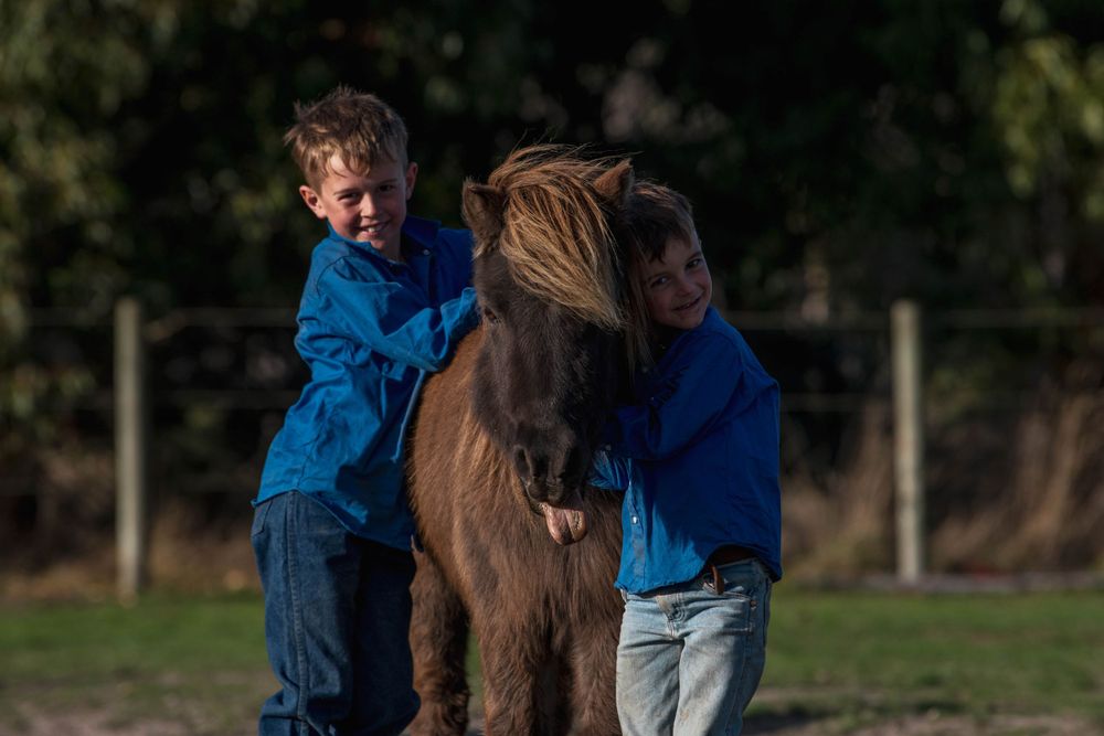 two children stand embracing their pony in a rustic countryside setting