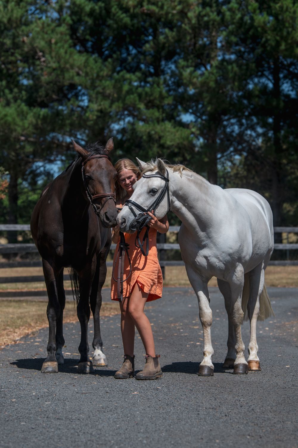 A person in an orange dress poses between a black and white horse on a paved surface.