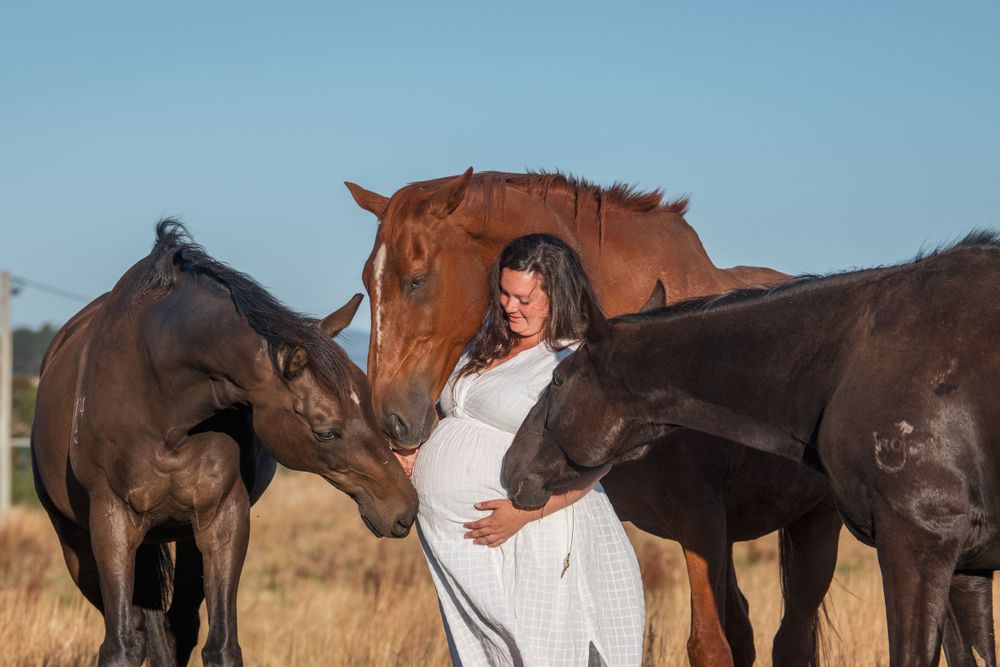 A person in a white dress poses with three dark horses in a rustic field setting.