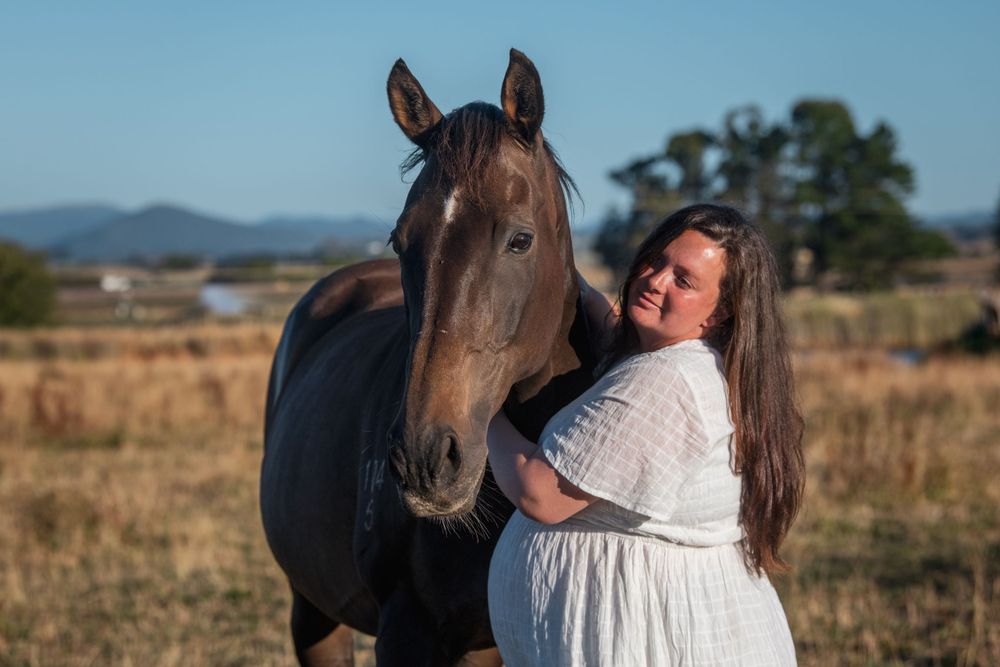 A person in a white dress embraces a dark horse in a field with mountains in the background.