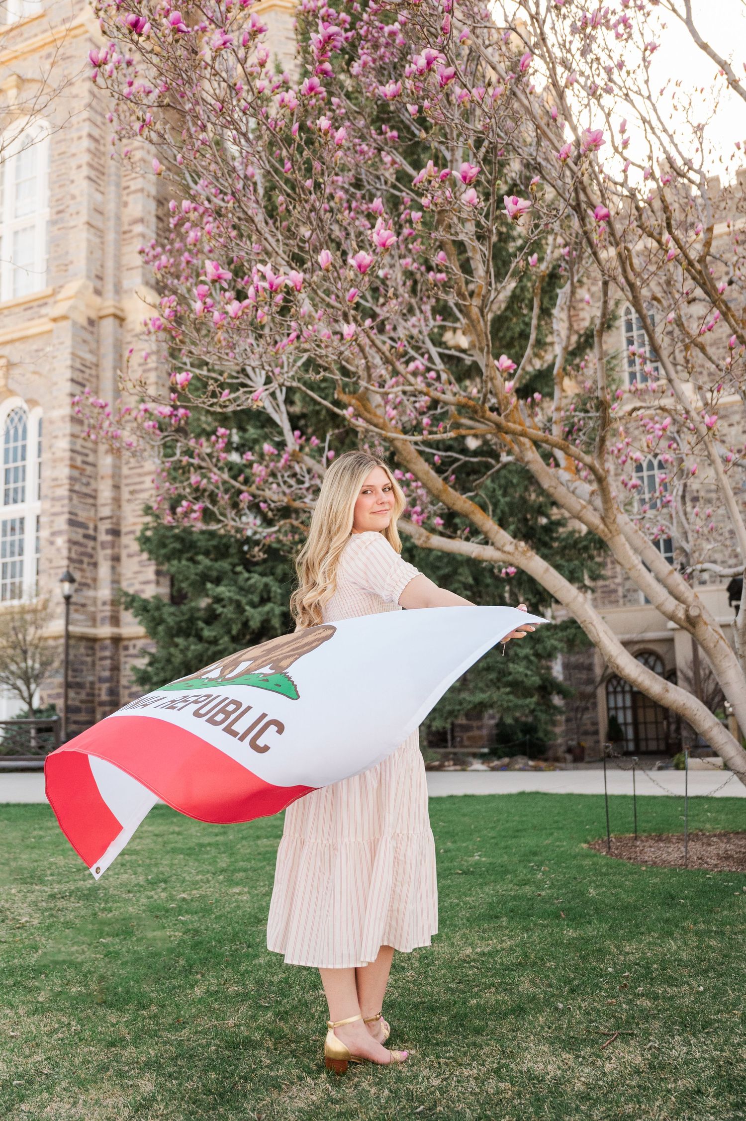 Person holding California flag in front of blooming magnolia tree and stone building.