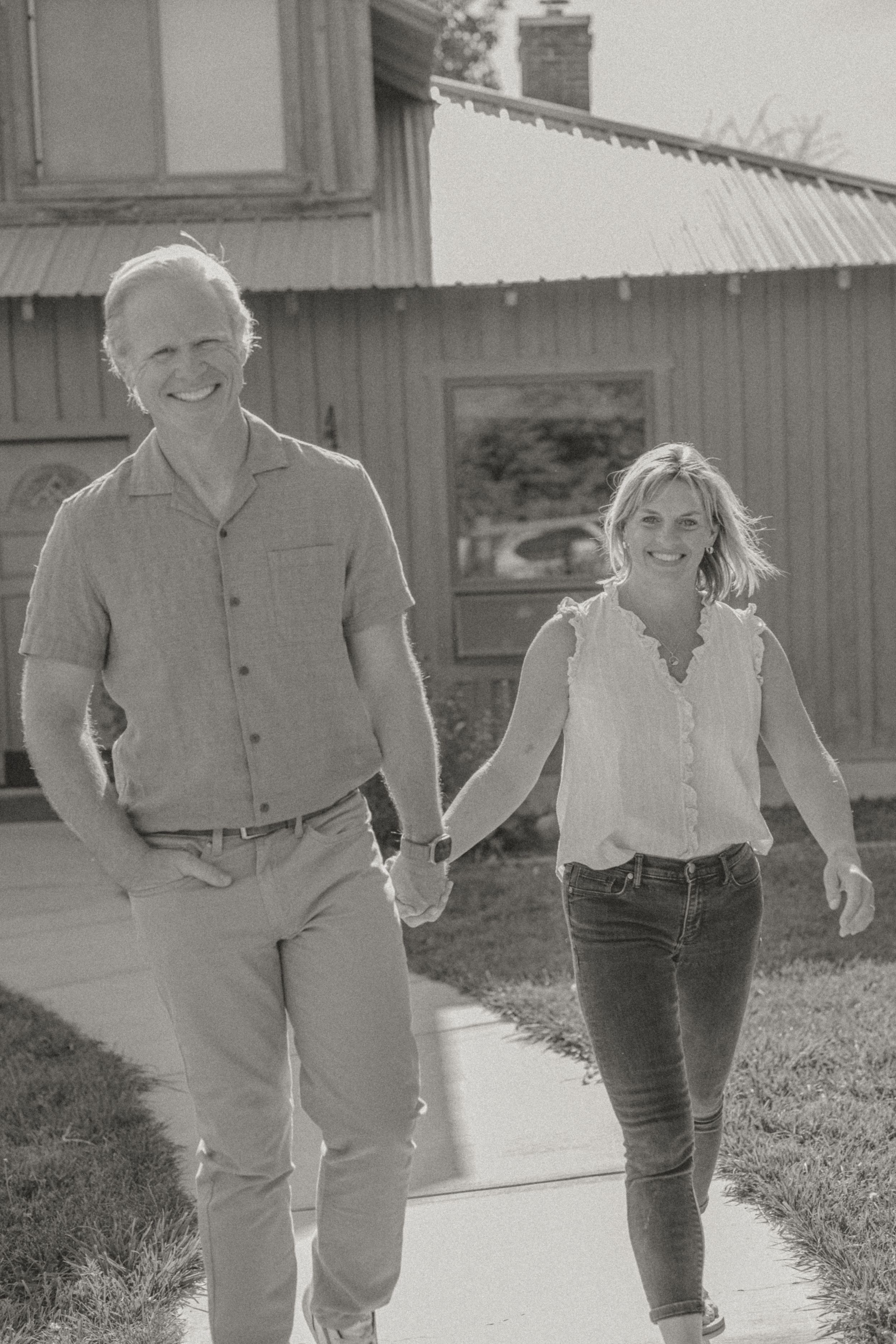 Black and white photo of a smiling couple holding hands while walking on a sidewalk outside a rustic building.