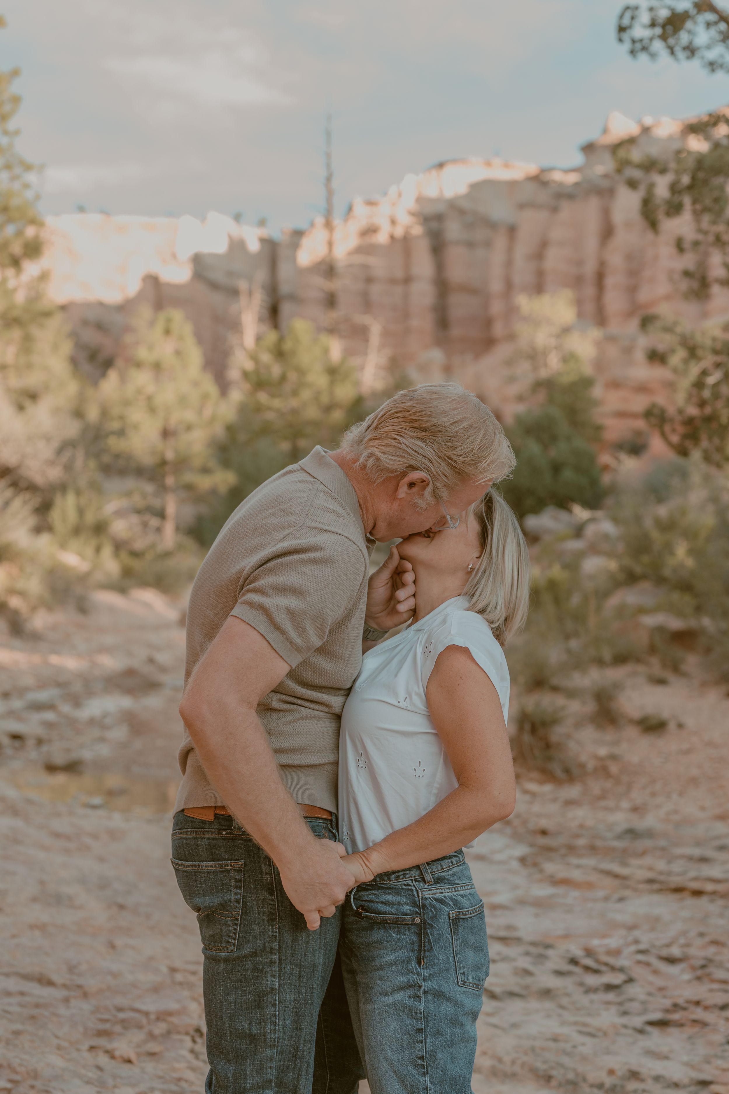 A couple shares a tender kiss surrounded by red rock canyon formations and desert landscape in warm golden light.