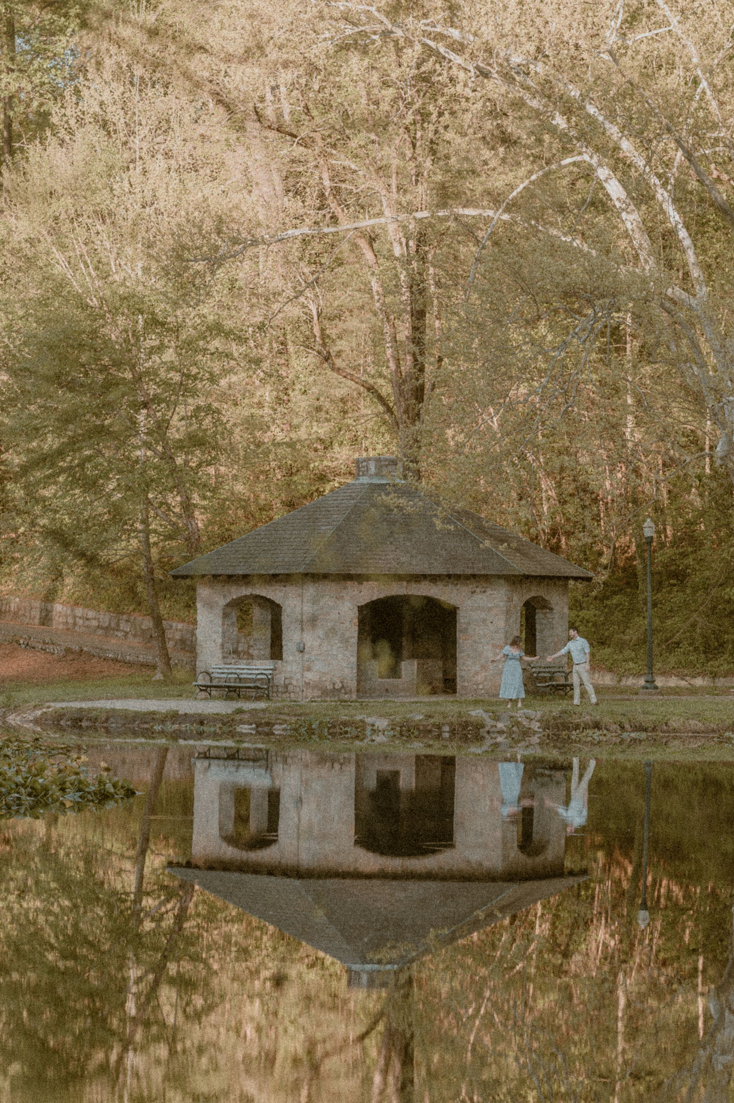 A rustic stone gazebo with multiple arched openings reflects perfectly in a still pond surrounded by autumn trees.