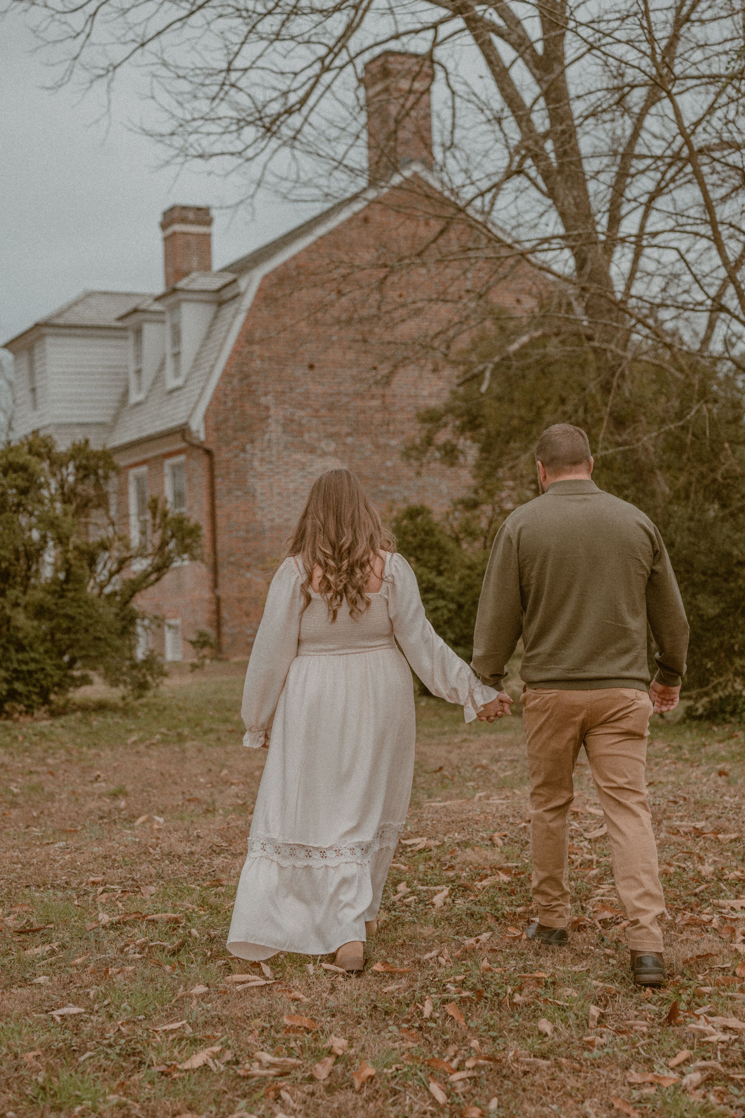 A couple holds hands walking toward a brick colonial house in autumn, woman in white dress, man in olive sweater.
