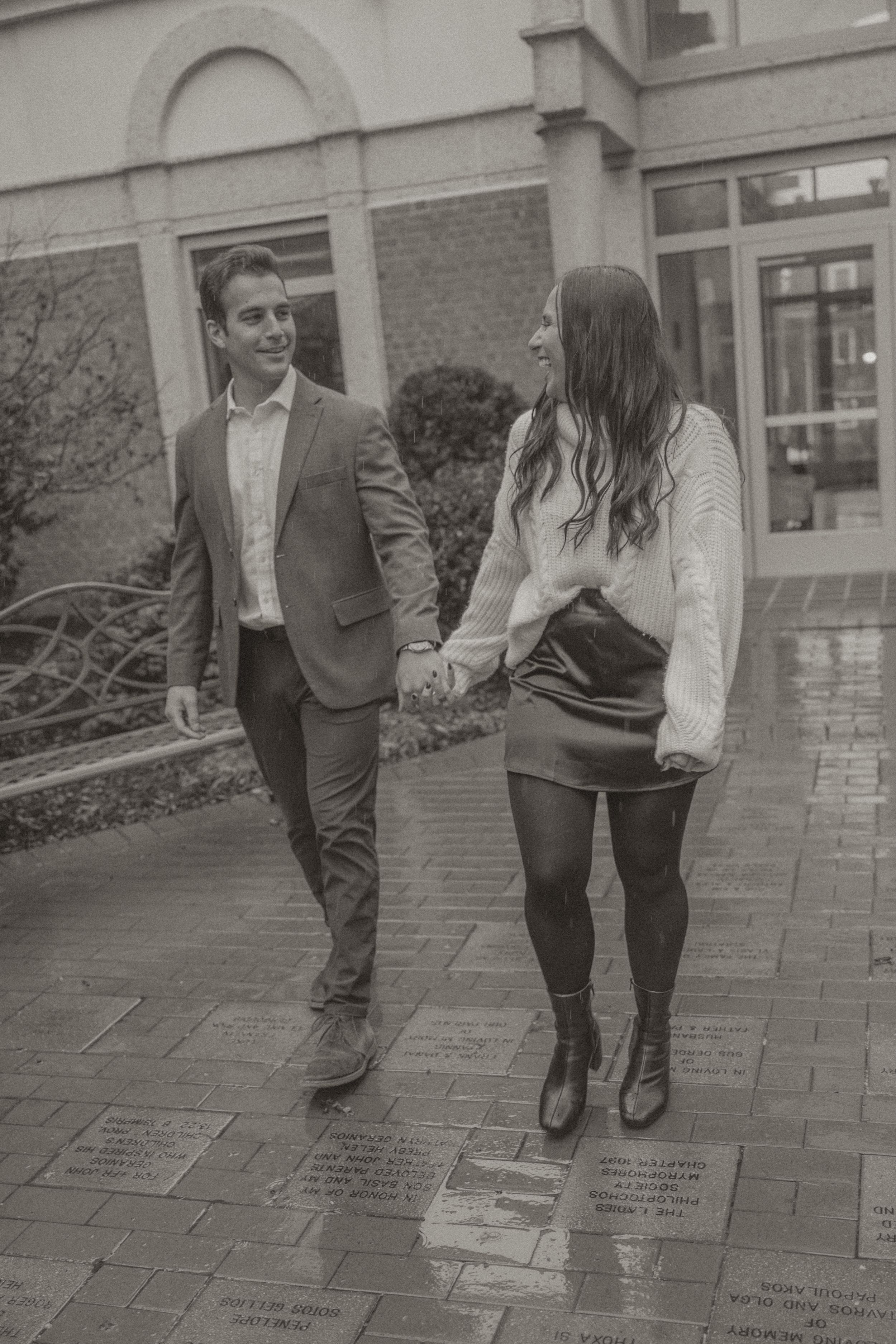 Couple holding hands walking on brick path in black and white photo, man in suit jacket, woman in mini skirt and boots.