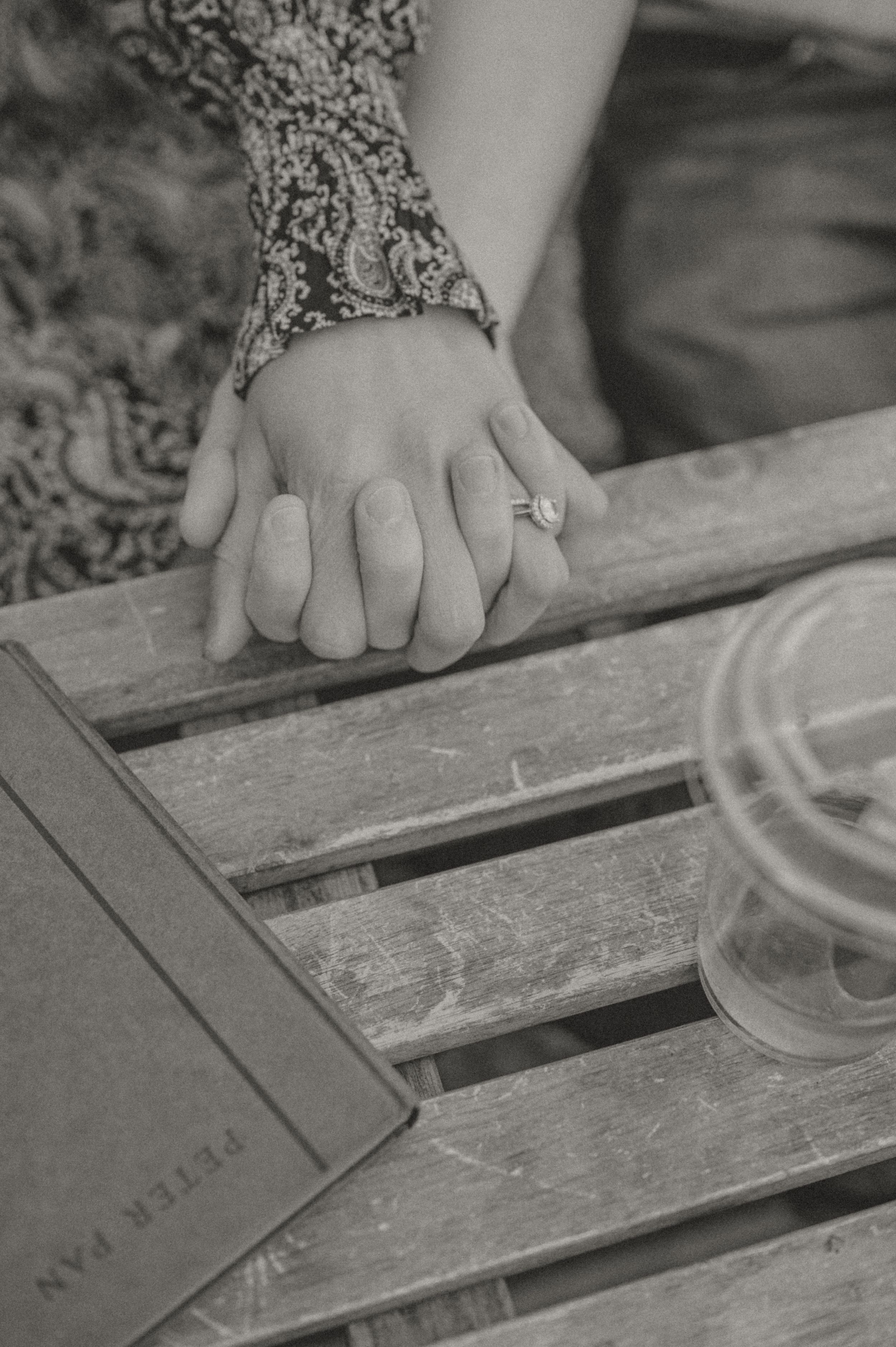 A hand with an engagement ring rests on a wooden table next to a glass and book in black and white.