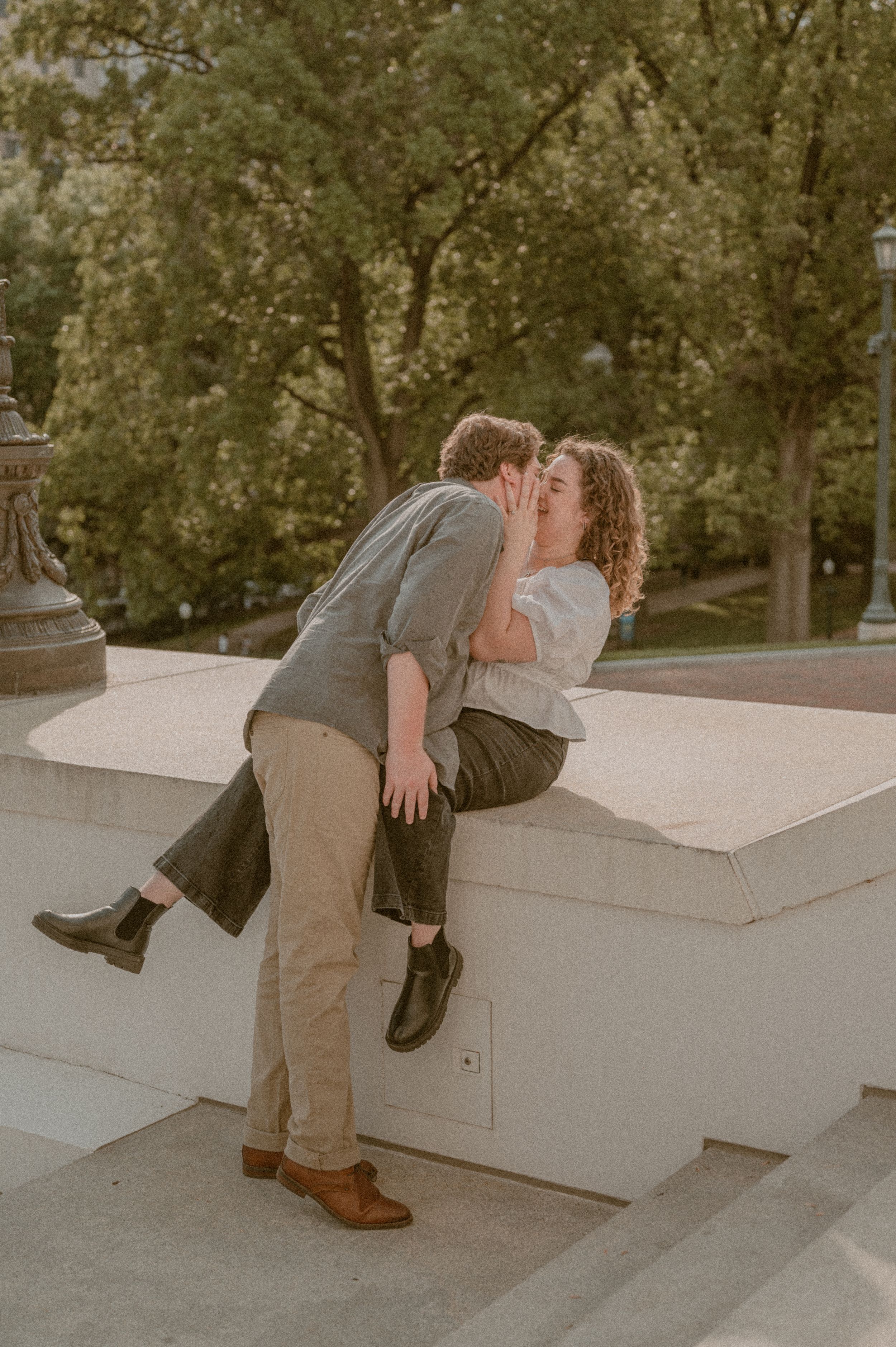 A romantic couple embraces on a white stone ledge in a park during golden hour.