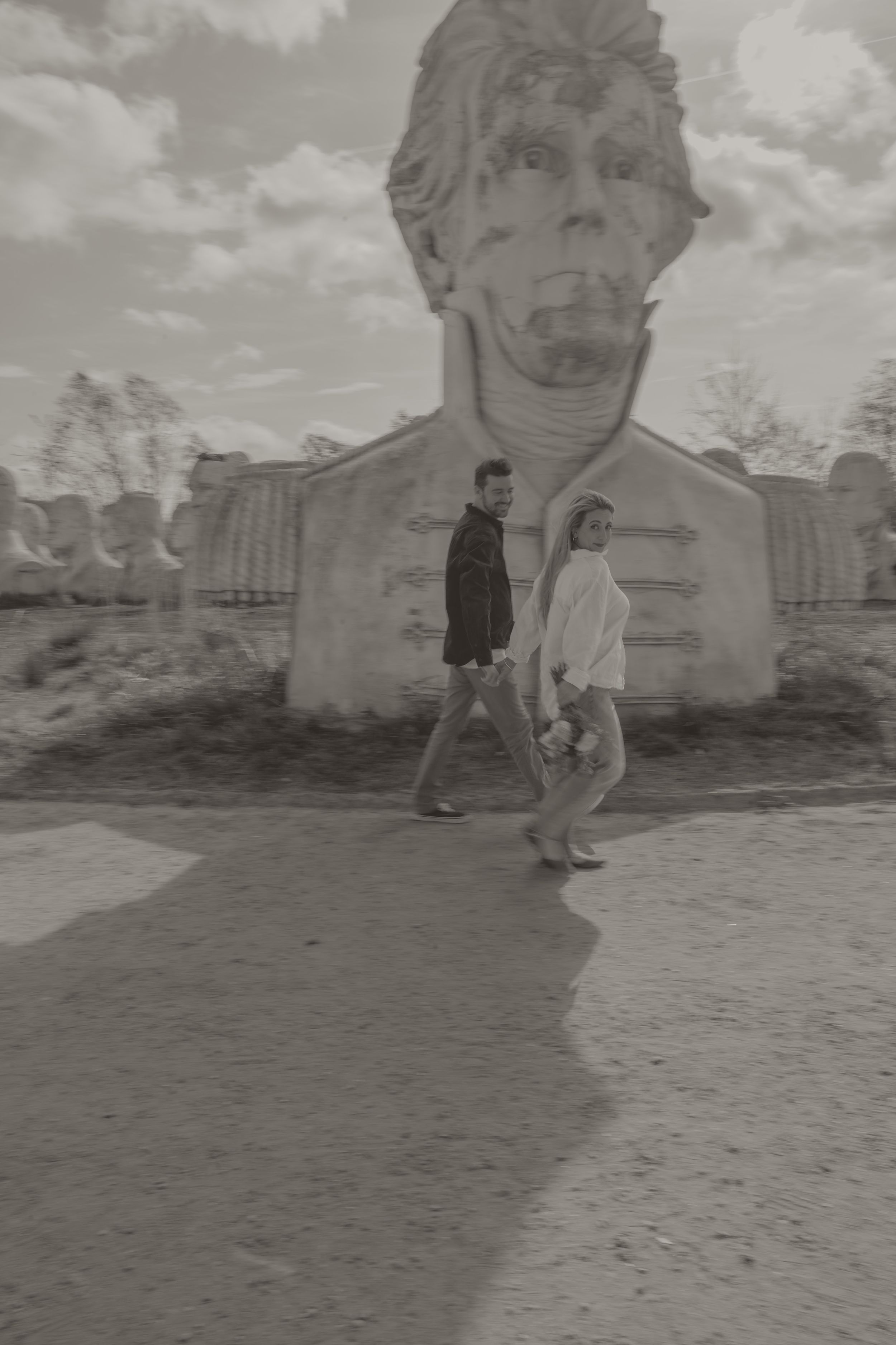 Black and white photo of a couple walking past a large stone face sculpture outdoors.