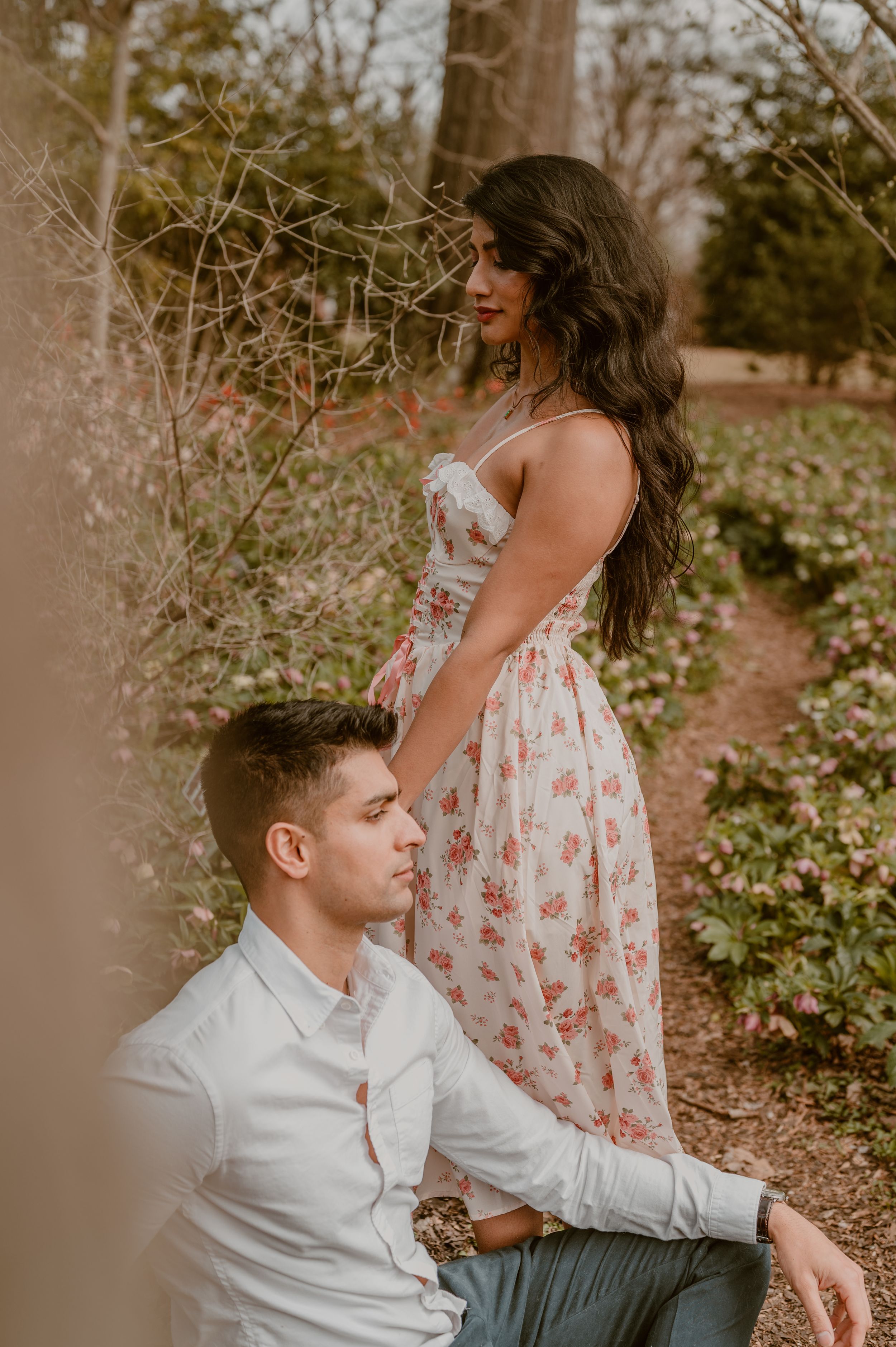 Romantic couple poses outdoors in natural setting with person in floral dress and person in white button-down shirt.