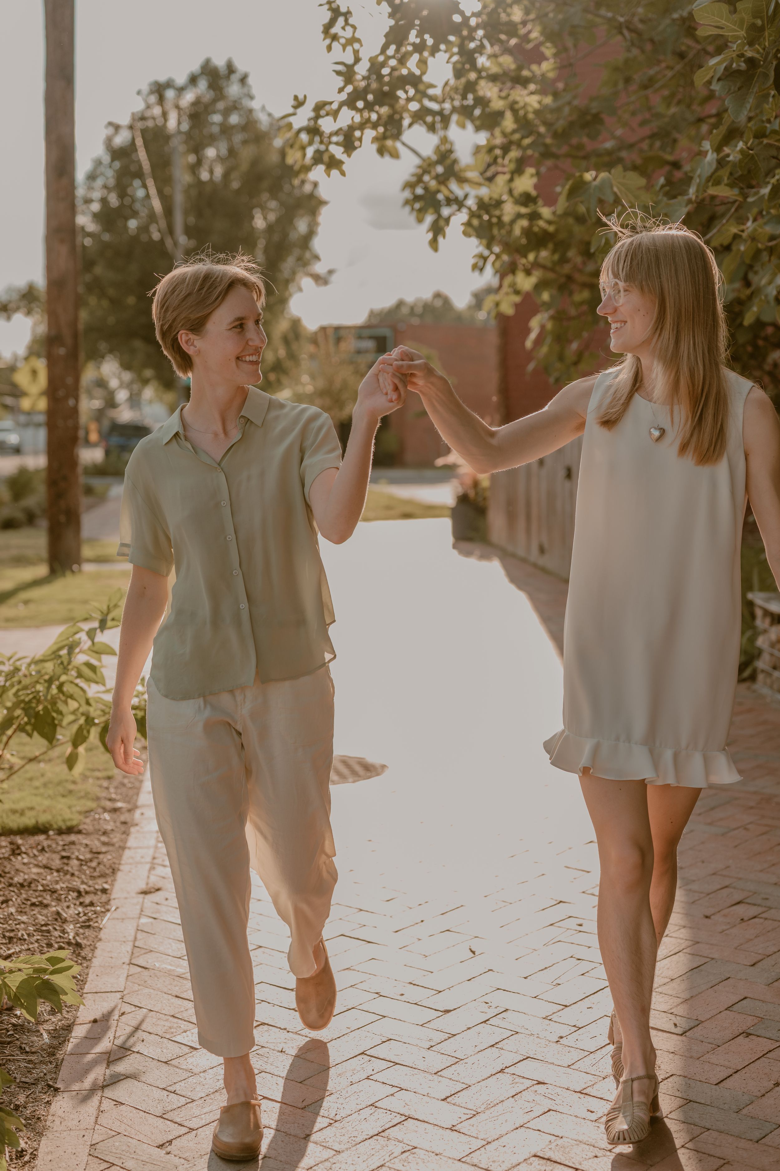 Two women holding hands and smiling while walking together on a brick path in warm sunlight.