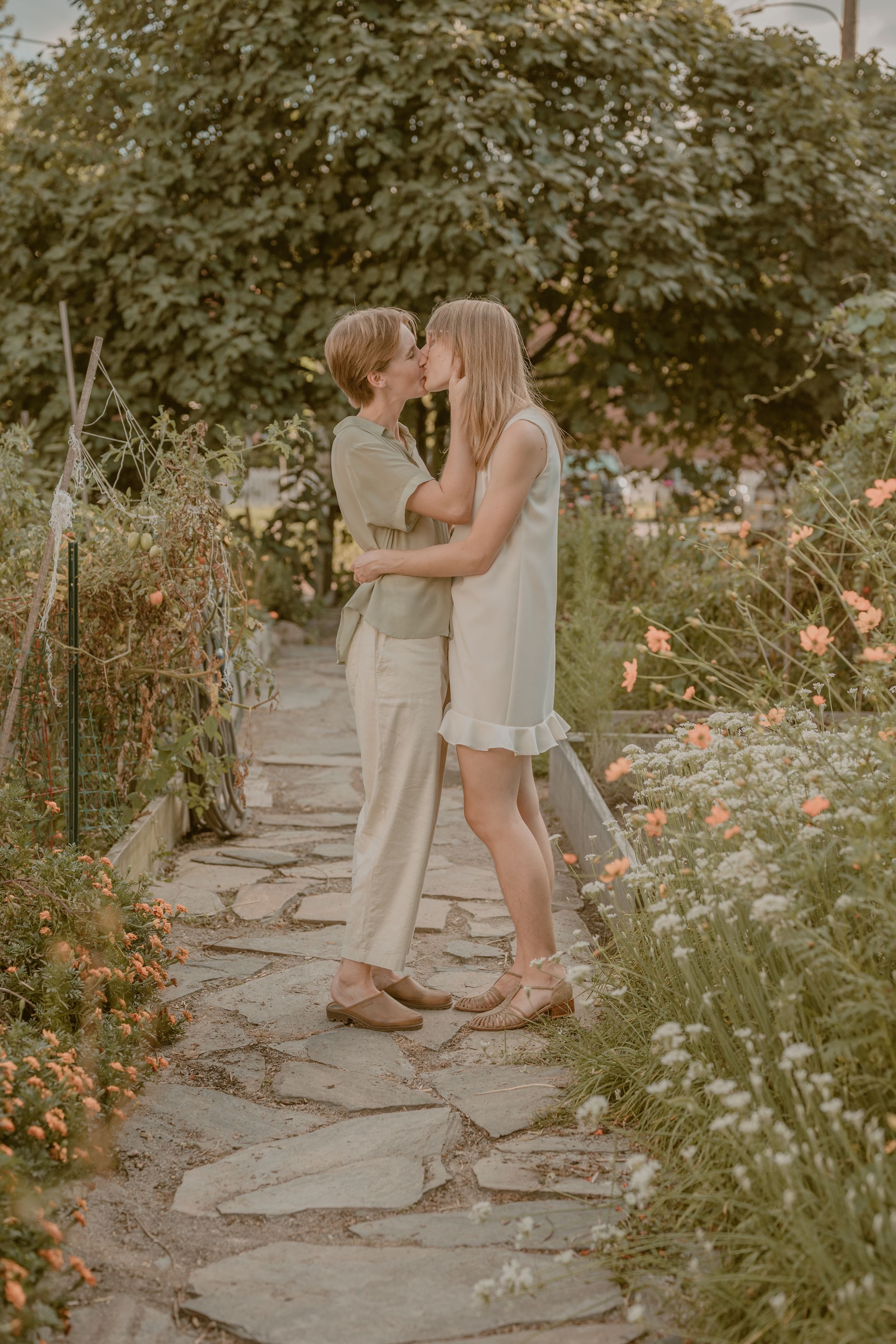 Couple sharing a romantic kiss on a stone garden path surrounded by blooming flowers and lush greenery.