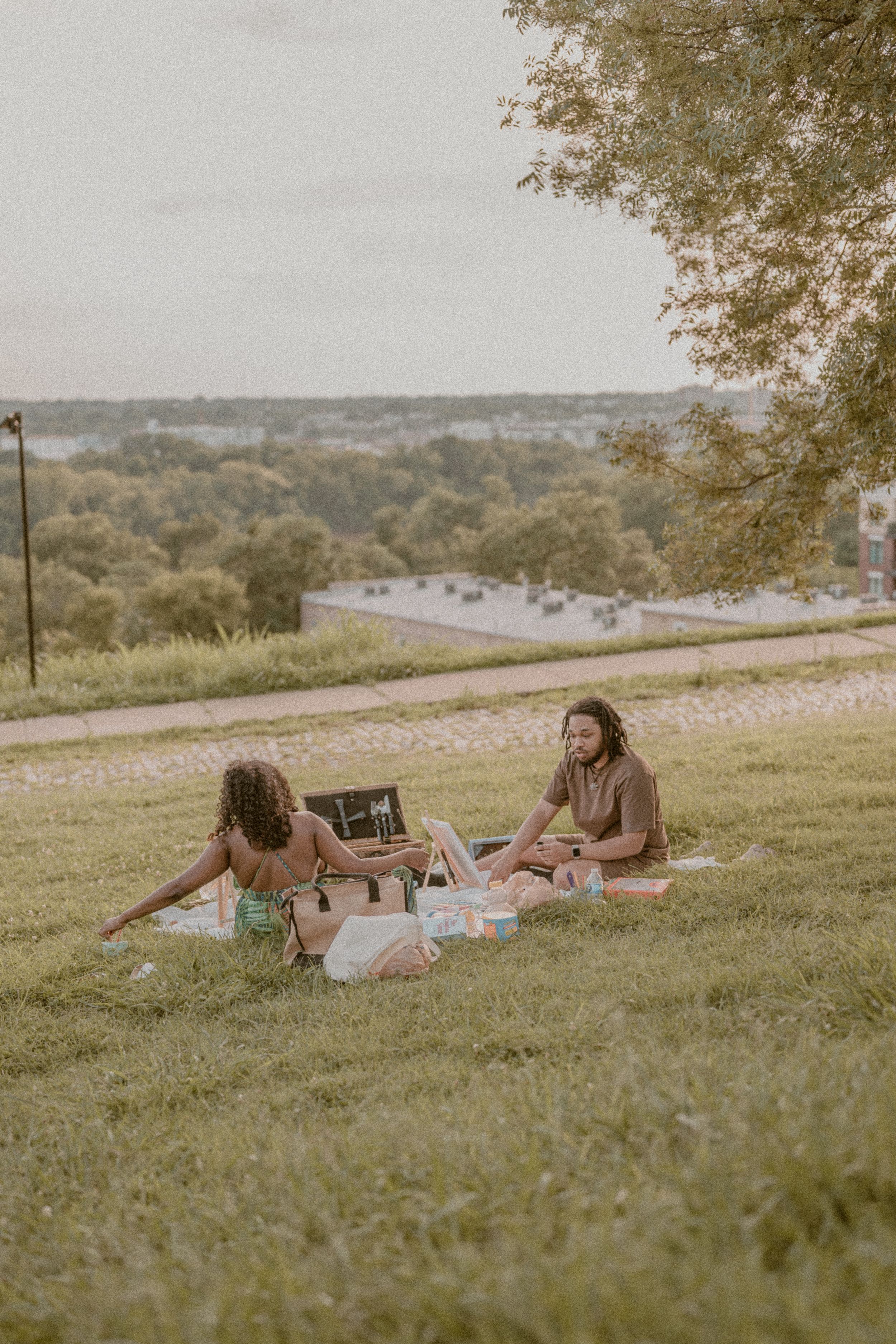 Two women enjoying a picnic on a grassy hill with scenic views of trees and a road in the background.
