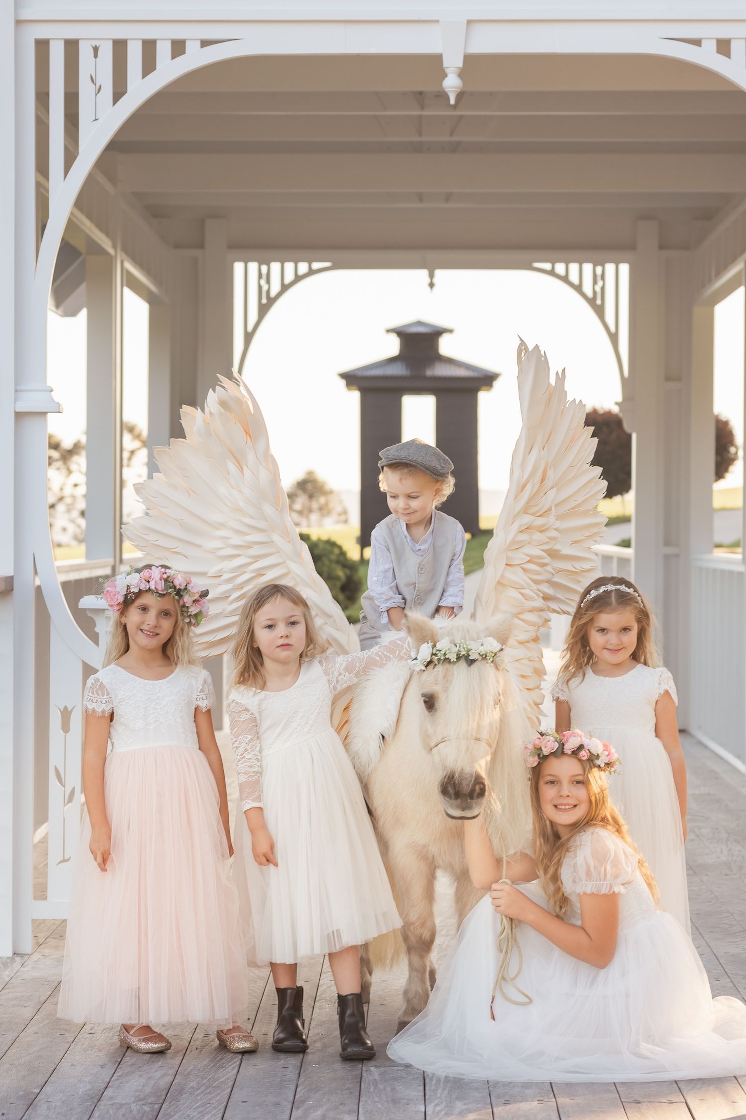 Group wearing white dresses and angel wings pose under a white gazebo.