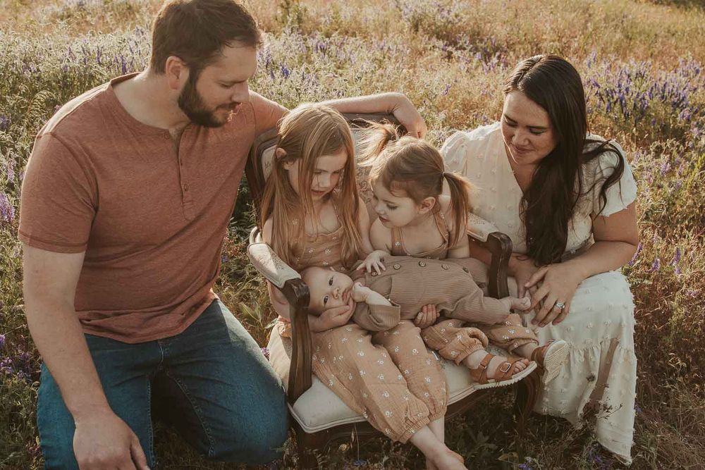 sisters sitting on vintage chair holding baby brother open flower field golden hour with a spokane family photographer