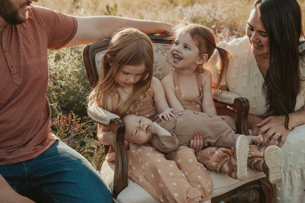 sisters sitting on vintage chair holding baby brother open flower field golden hour with a spokane family photographer