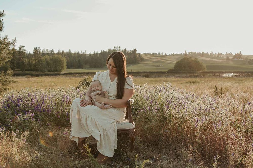 mother dressed in white dress holding baby while sitting in a chair in flower field in spokane