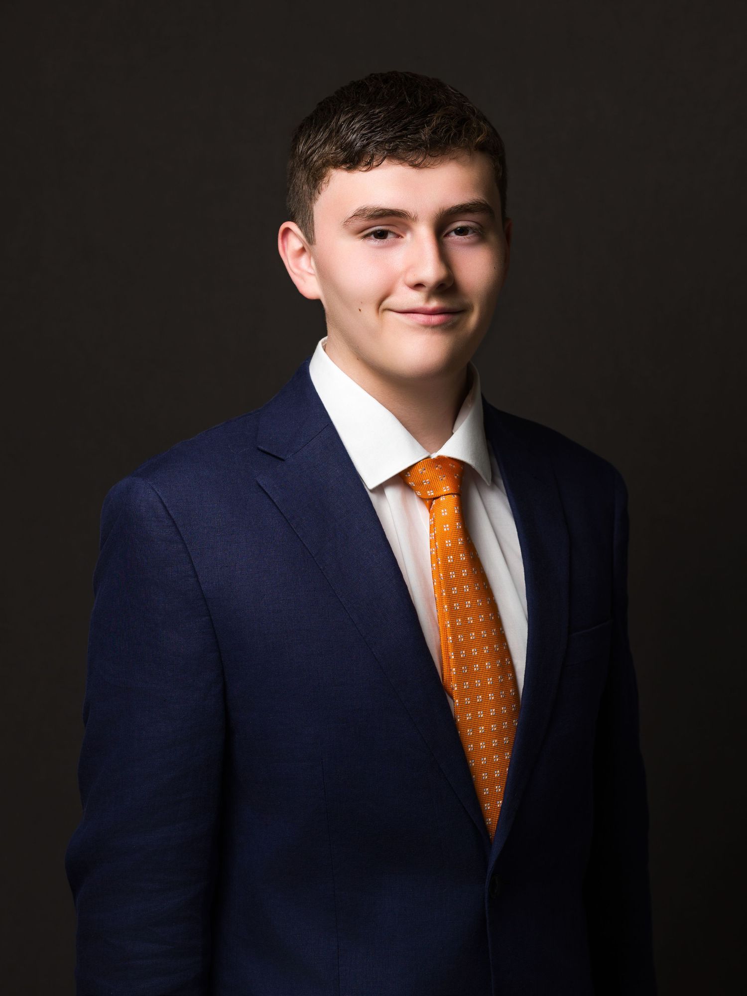 Professional headshot portrait of someone wearing a navy suit and orange polka dot tie against a dark background.
