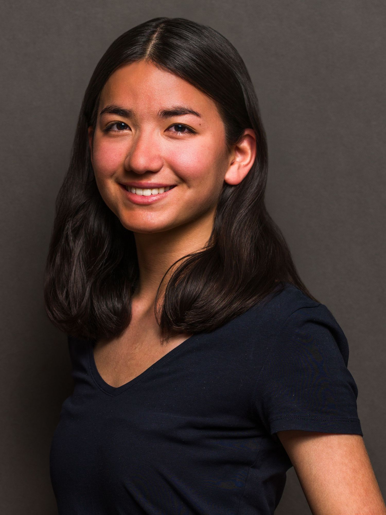 Young woman with dark hair smiling confidently in a navy v-neck top against a neutral gray background.