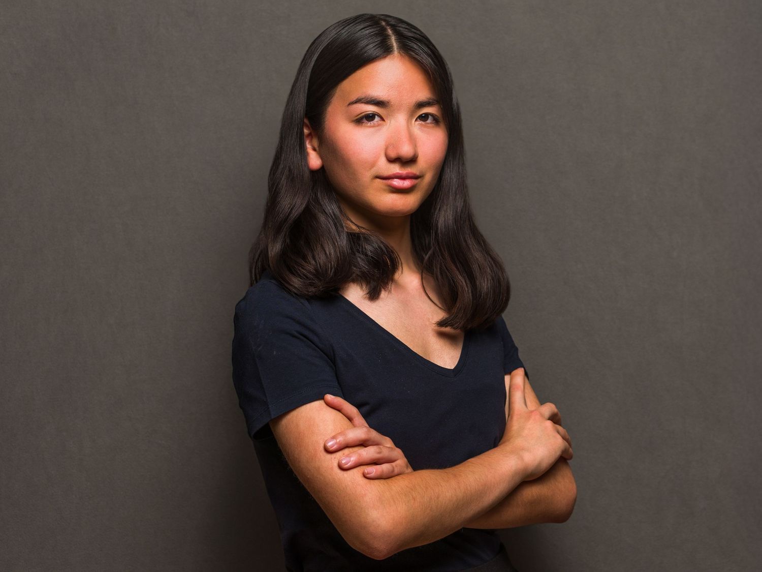 Young woman with crossed arms in navy top poses confidently against gray background.