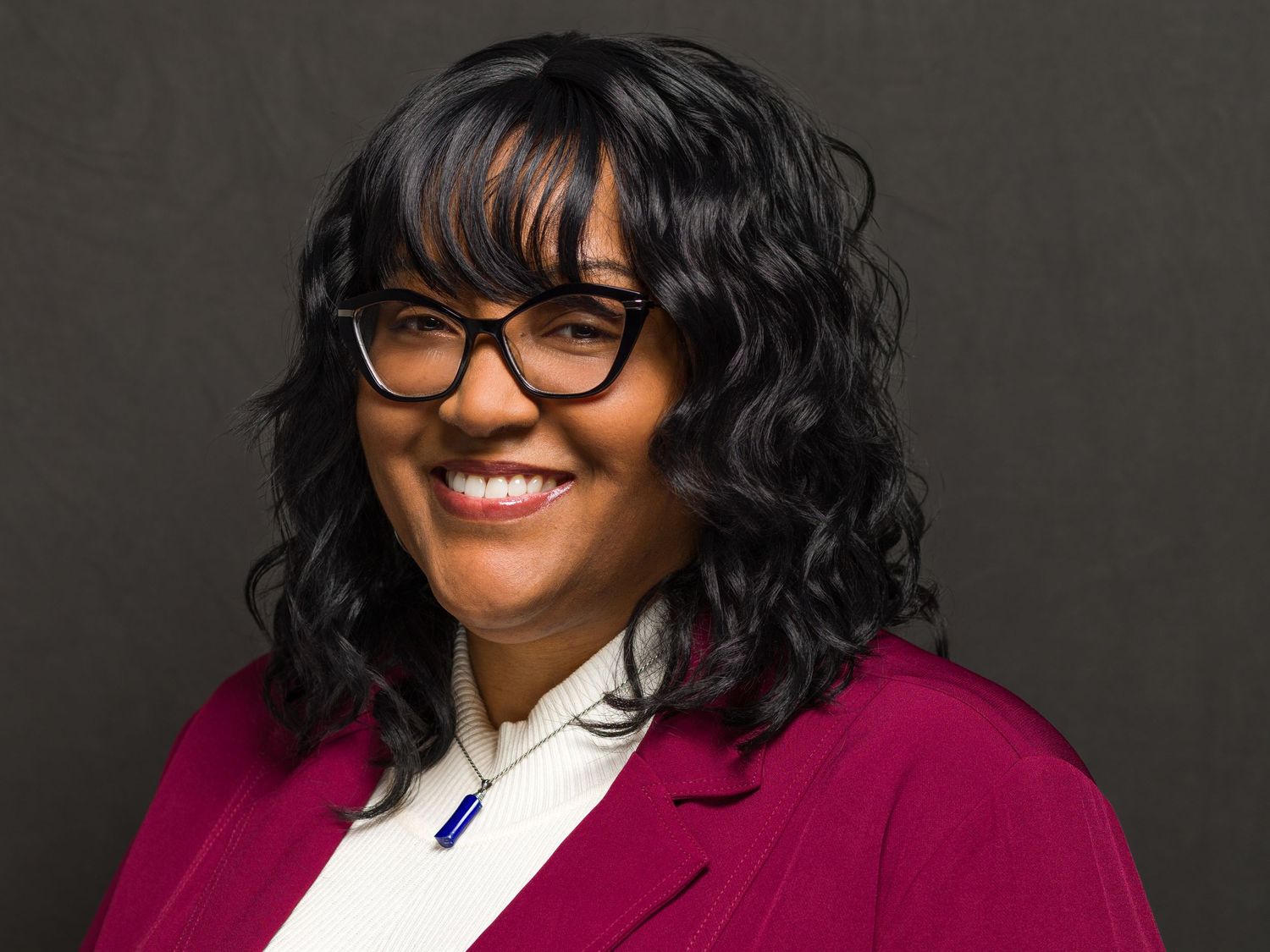 Studio headshot of someone in magenta blazer and white shirt with black rimmed glasses.