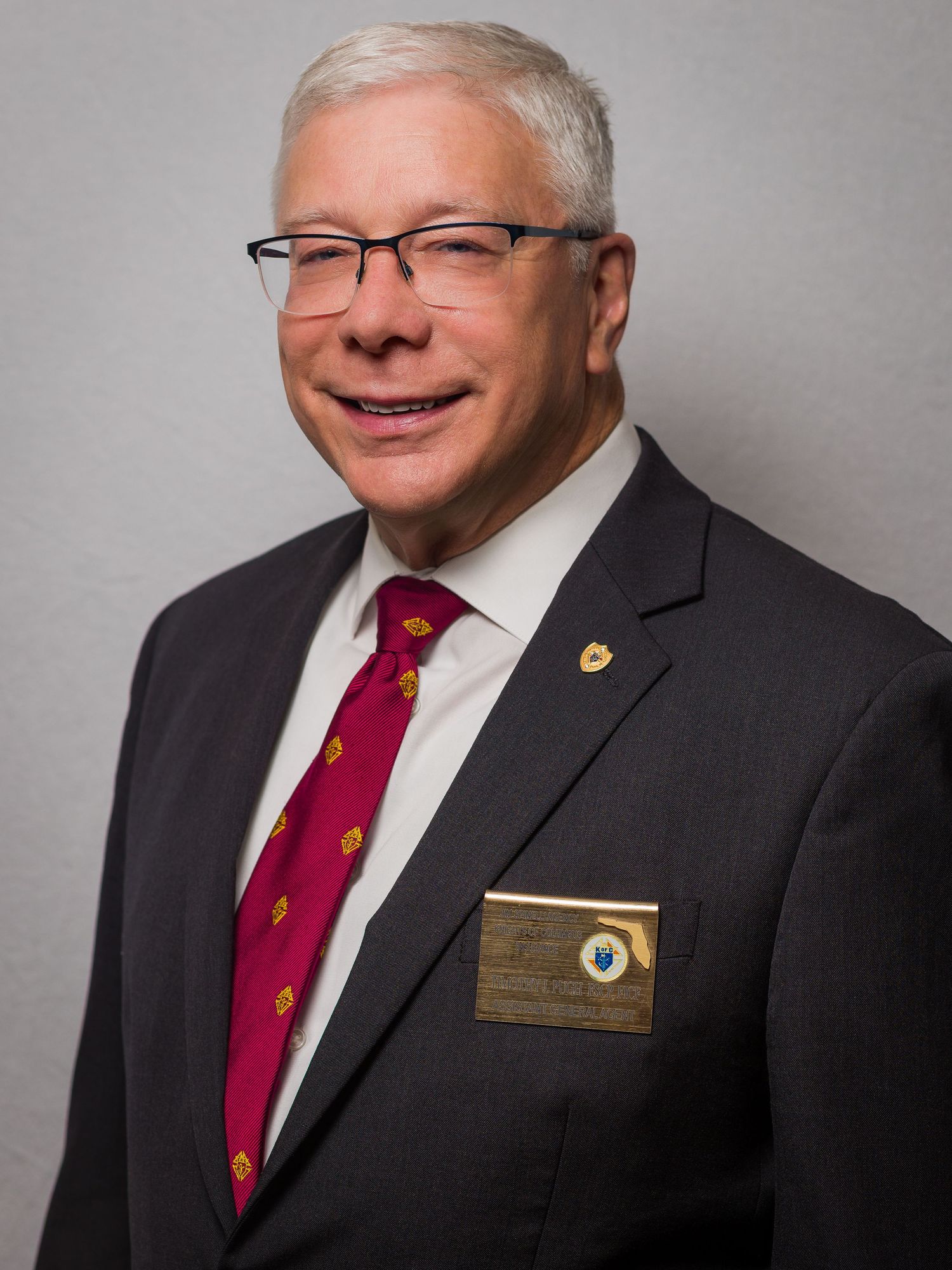 Professional headshot of a person wearing glasses, a dark suit jacket and red patterned tie with a name badge.