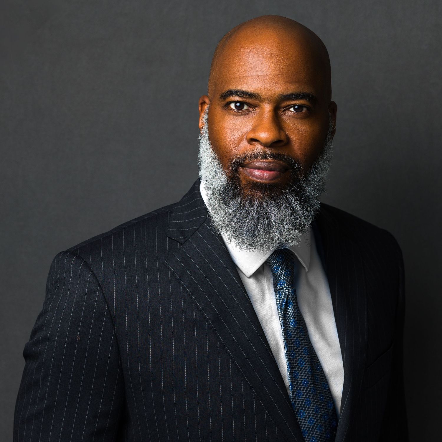 Professional Black man with gray beard wearing pinstripe suit and tie poses for formal business portrait.