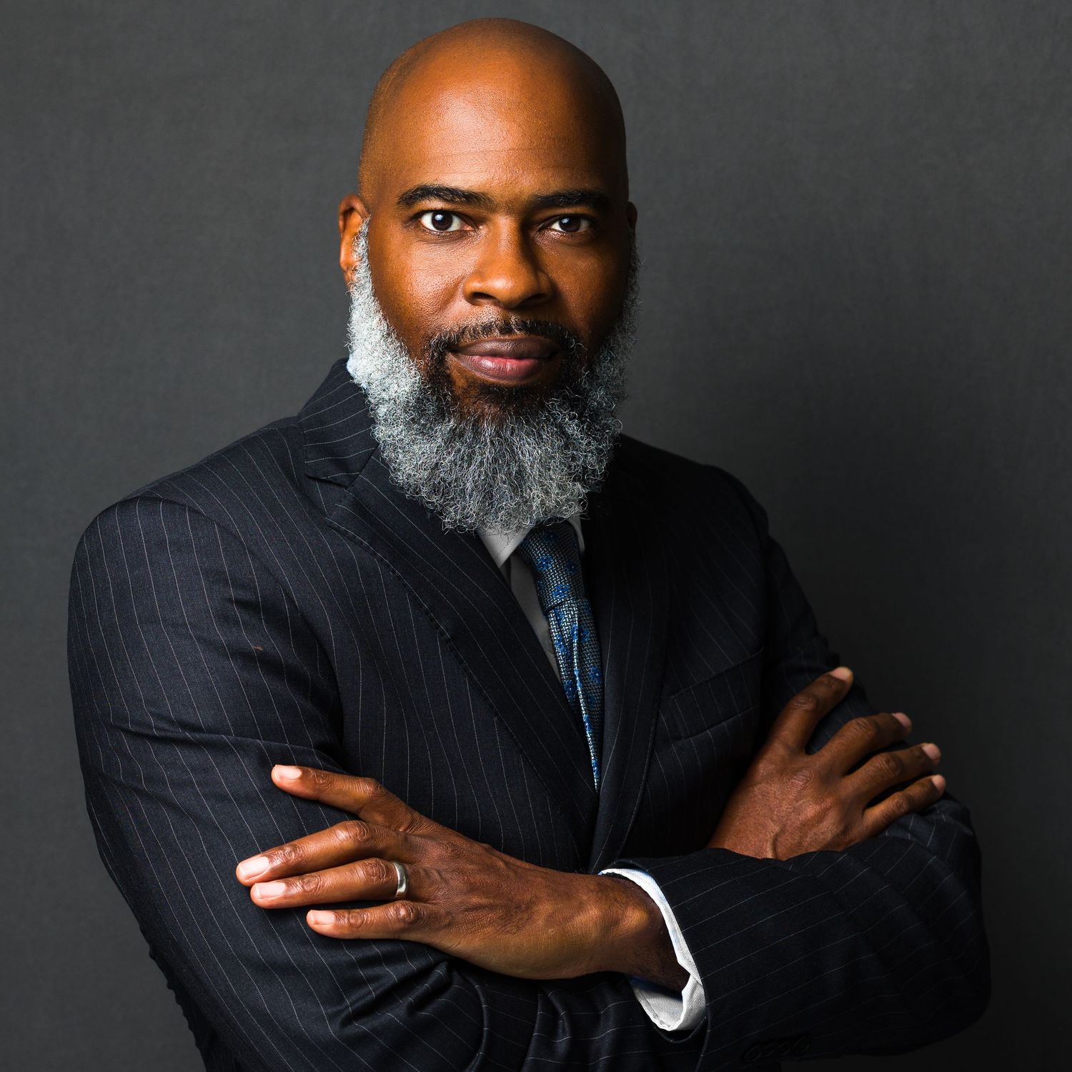 Professional Black man with gray beard in pinstripe suit, arms crossed, posing confidently against gray background.
