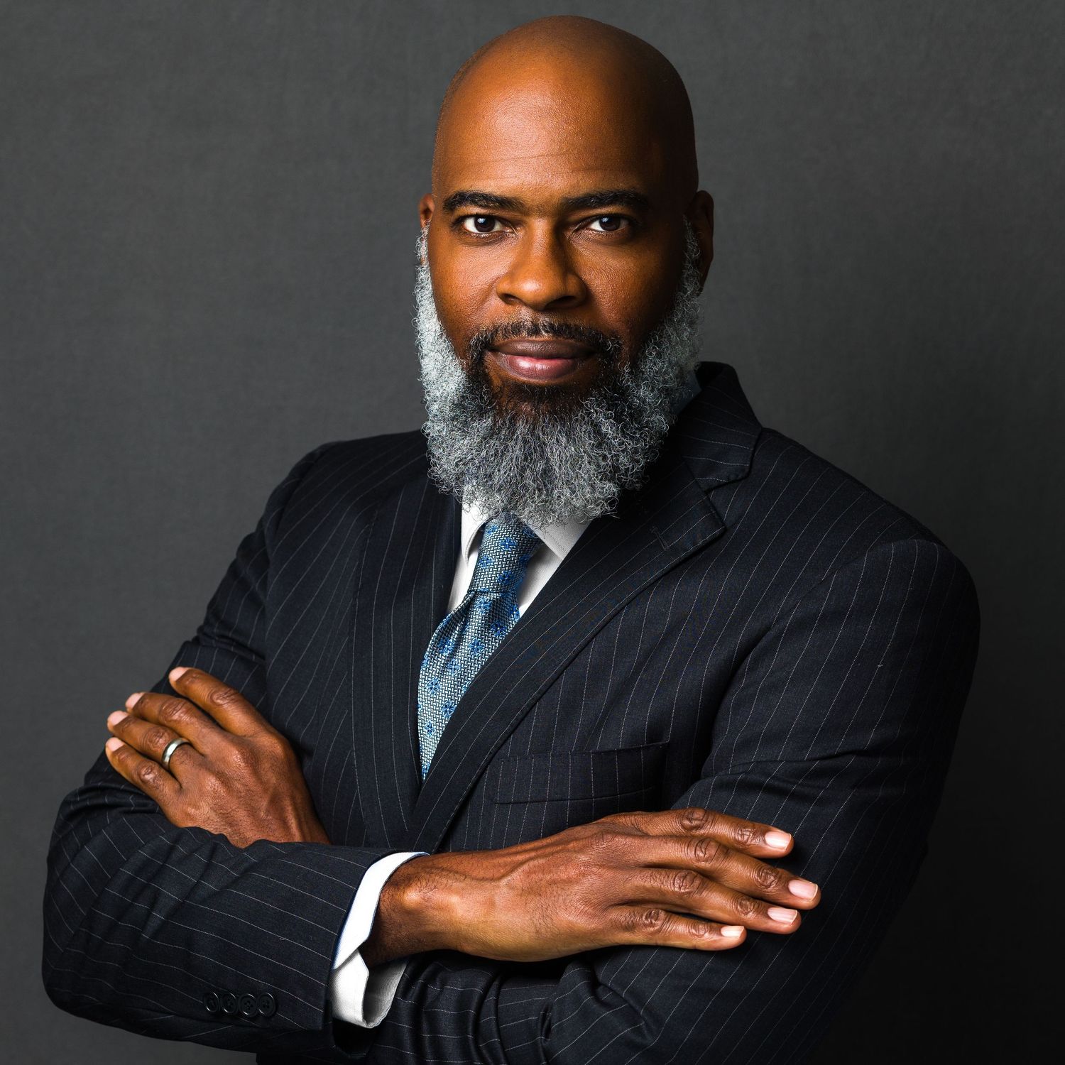 Professional Black man in pinstripe suit with gray beard, arms crossed, posing confidently against dark gray background.