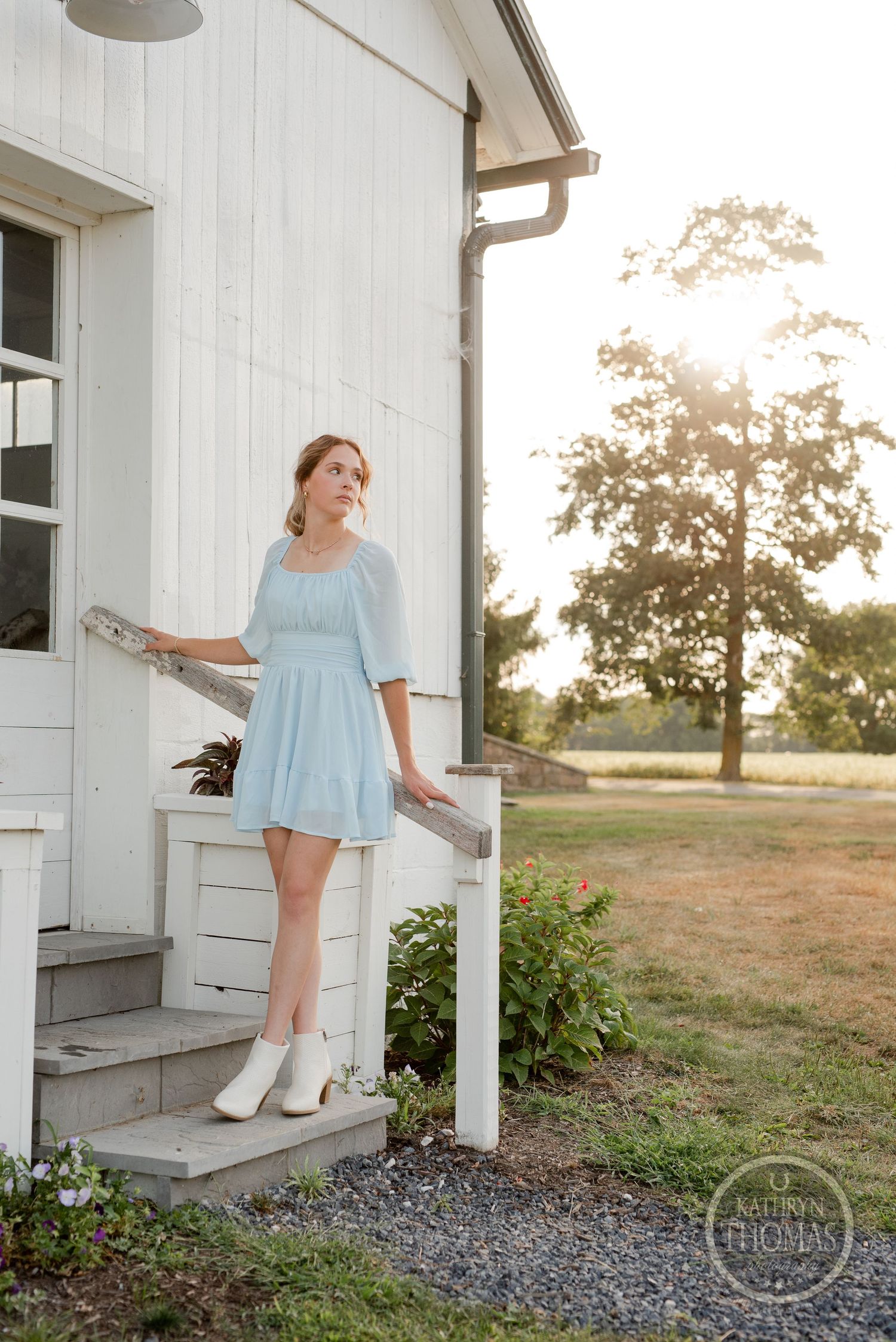 Light blue dress contrasts against classic white farmhouse exterior in golden evening light.