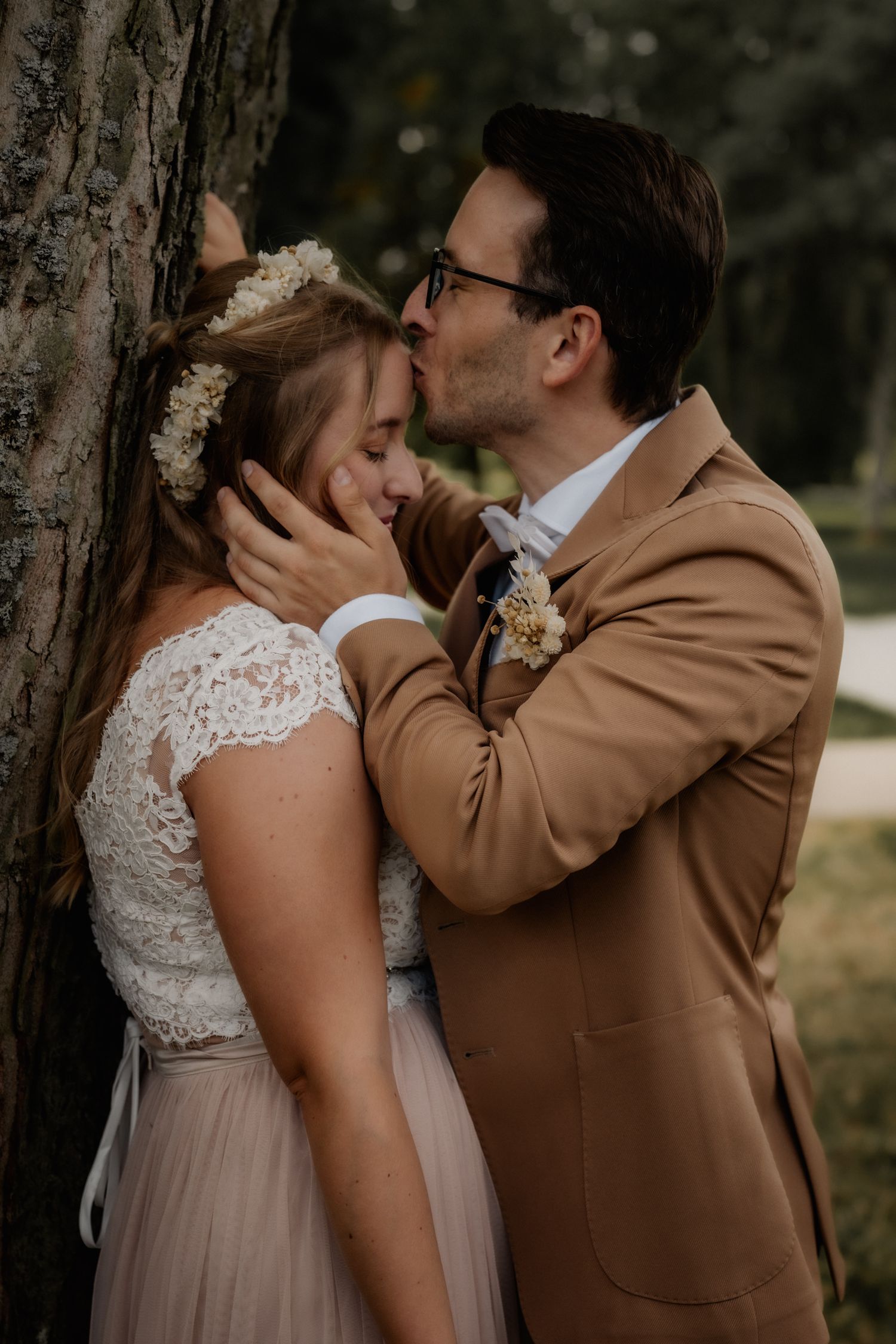 Romantic wedding moment shared between couple in formal attire against tree trunk.