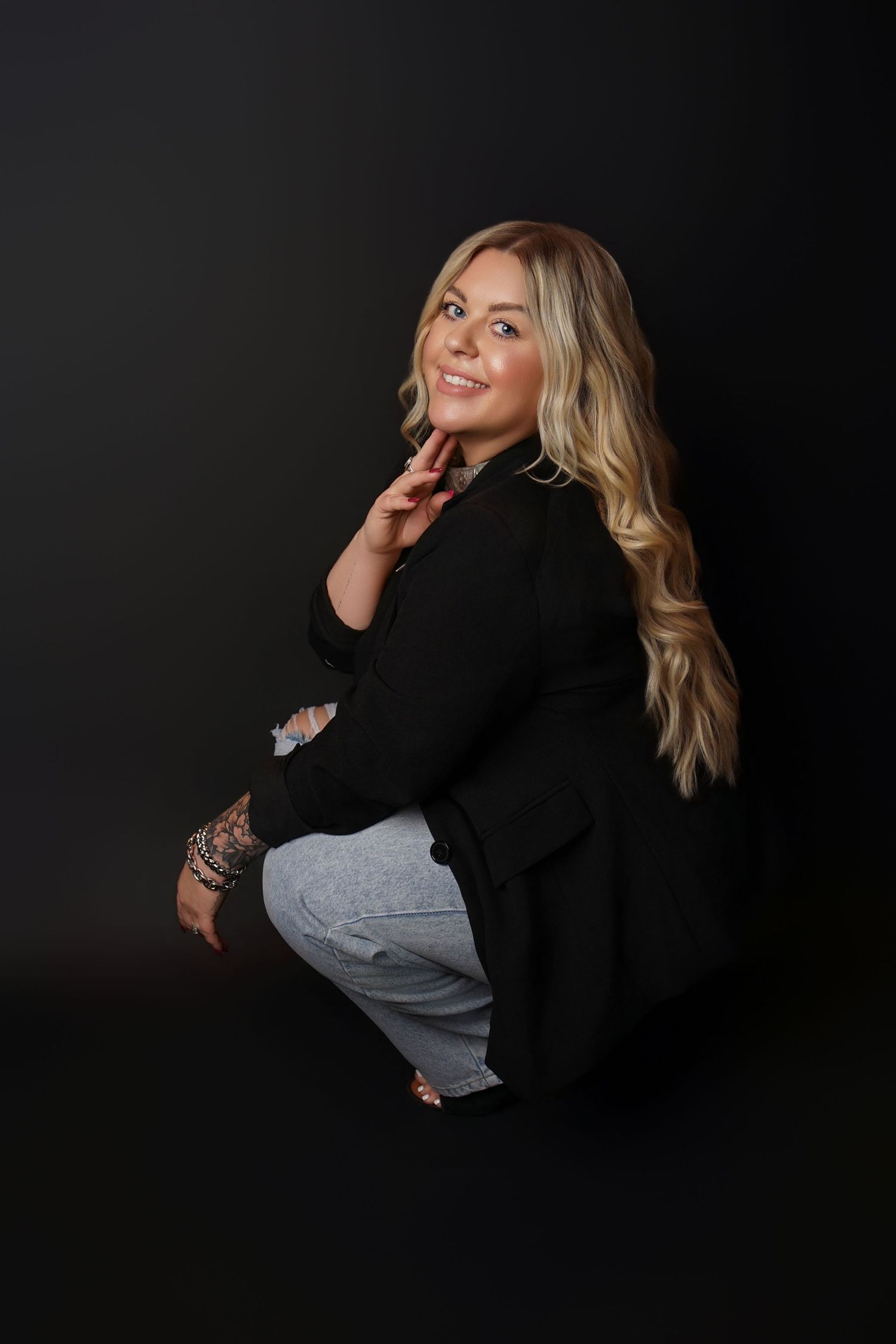 Smiling woman with long wavy blonde hair in a black blazer and light jeans crouching against a dark studio backdrop.