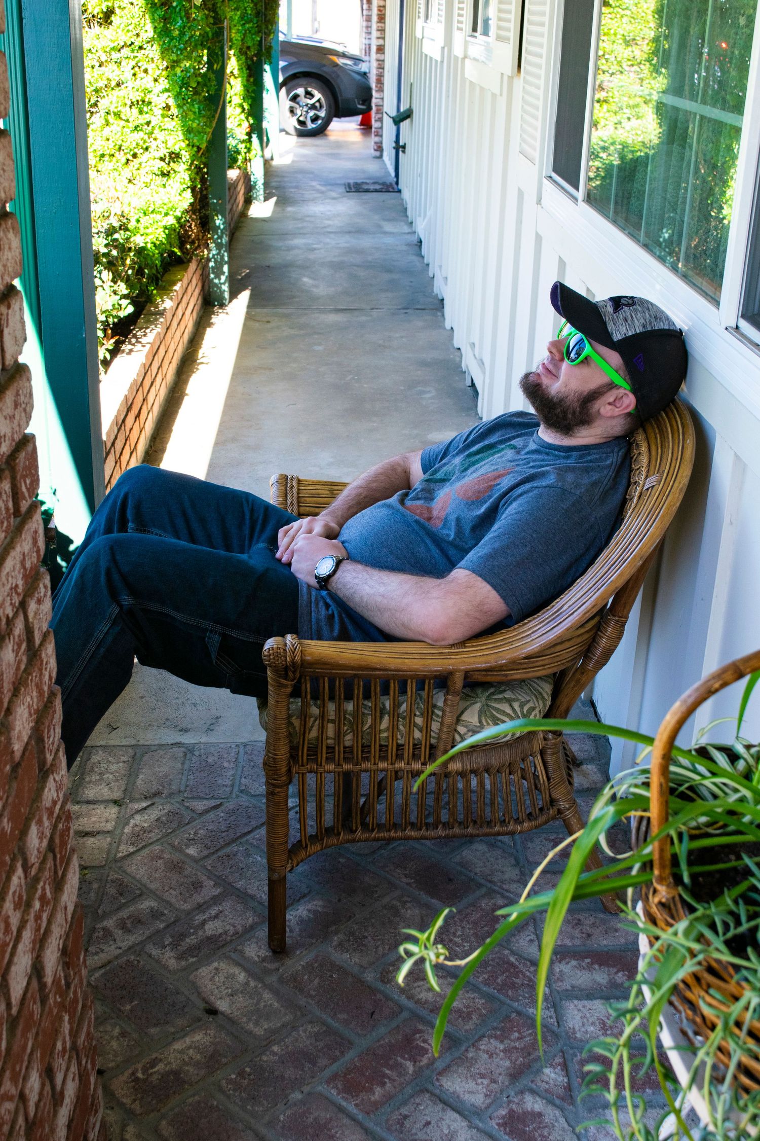 A person relaxing in a wicker chair on a narrow porch with plants.