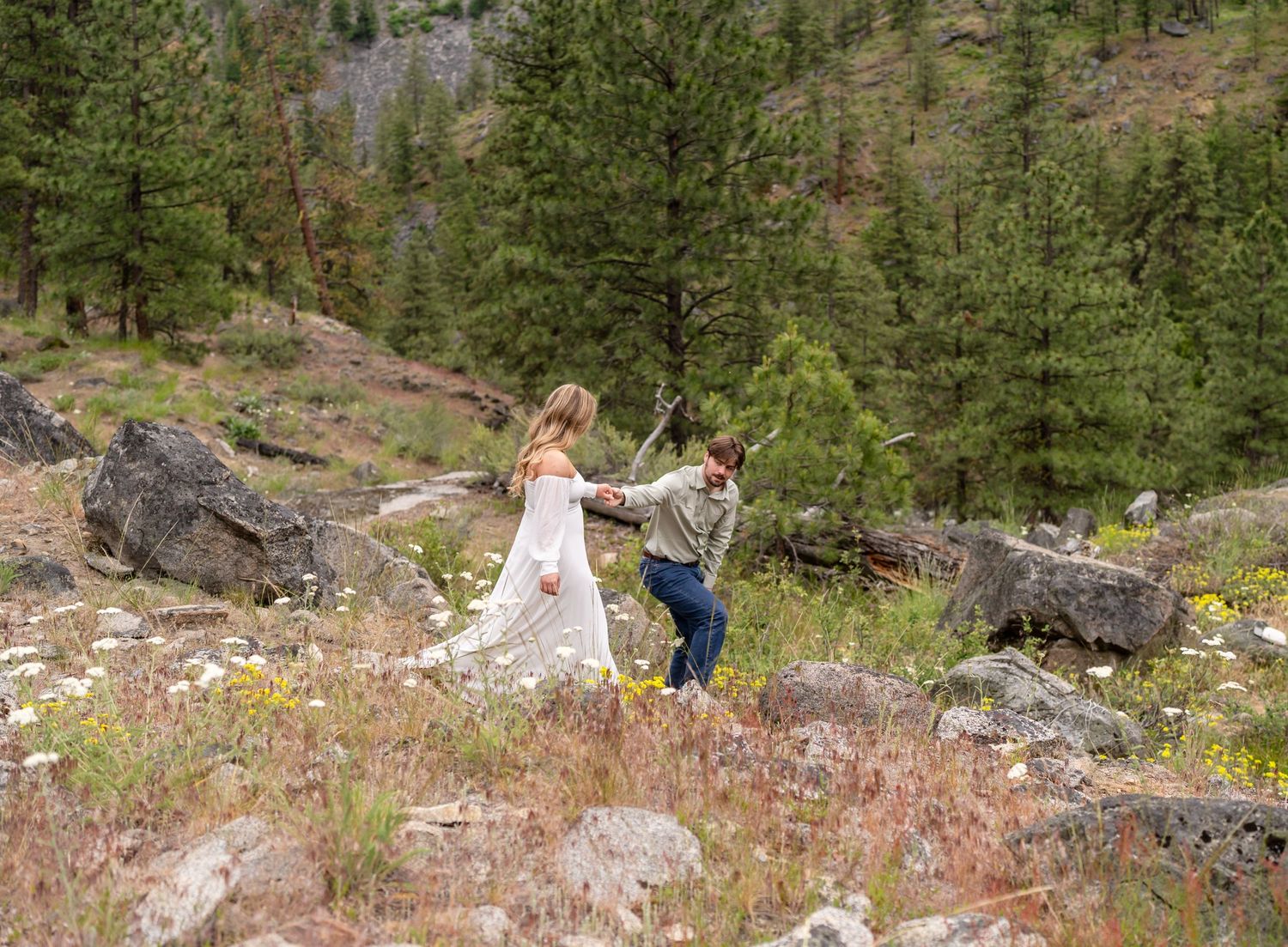 A couple shares a joyful moment on a rocky mountain slope surrounded by pine trees.