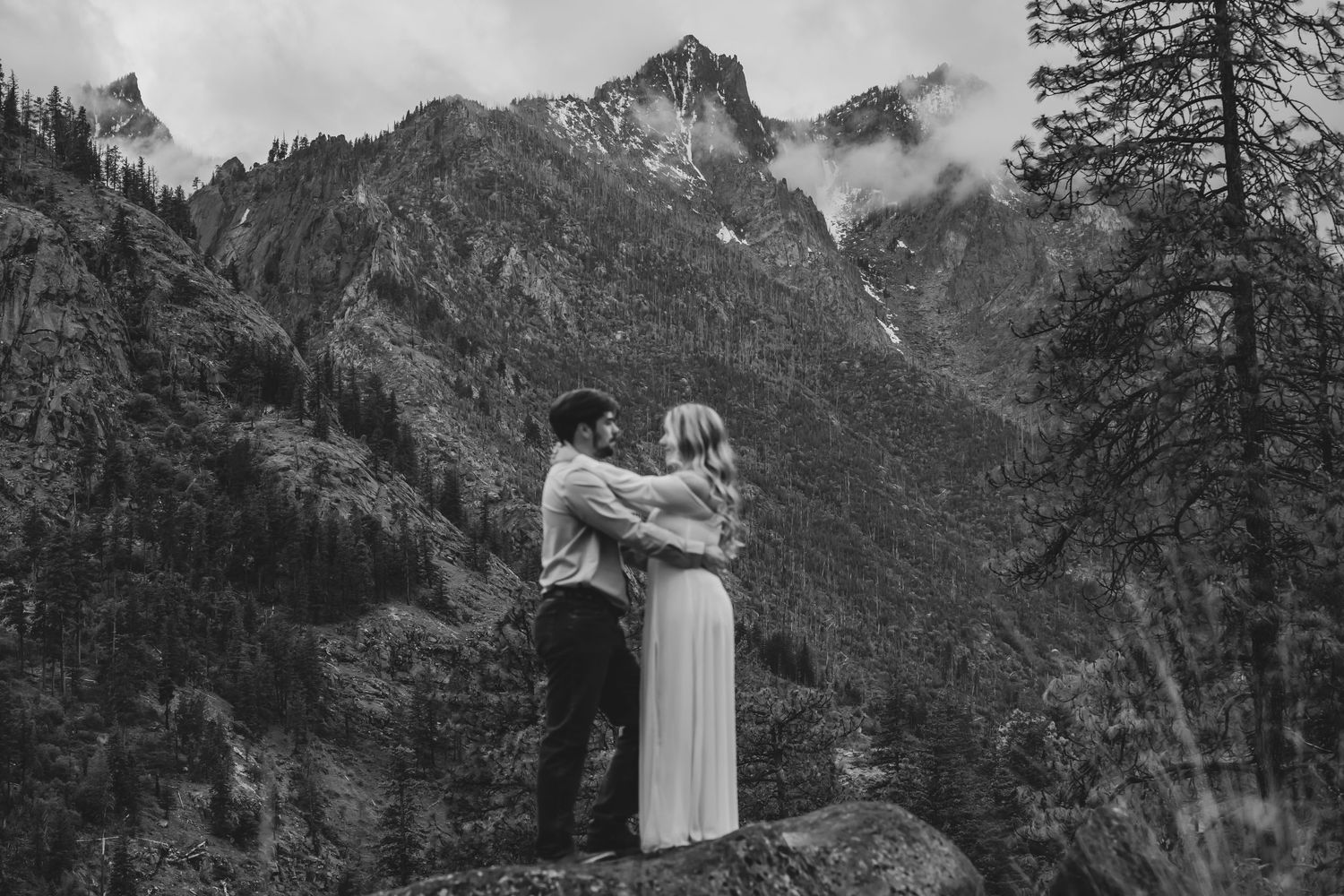 A couple embraces against dramatic mountain peaks in a moody black and white landscape.