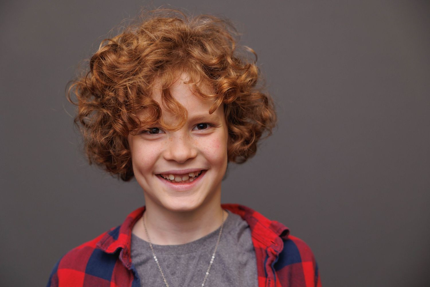 Portrait of a child with curly red hair wearing a red and gray plaid shirt against a dark background.