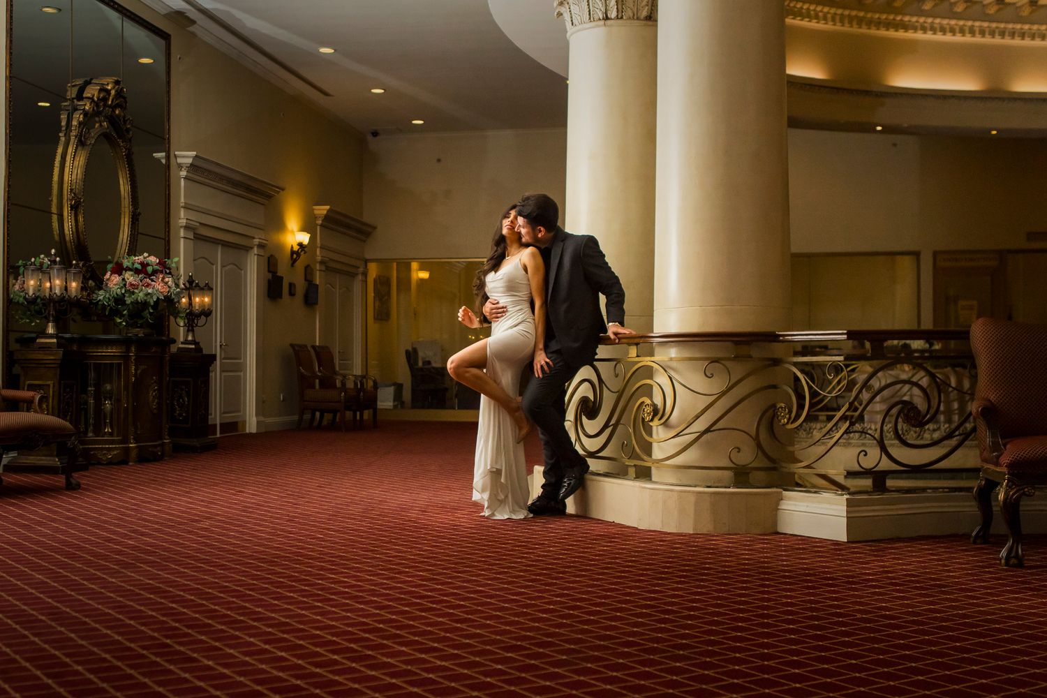 Romantic couple posing by an ornate column in an elegant hotel lobby with red carpeting.