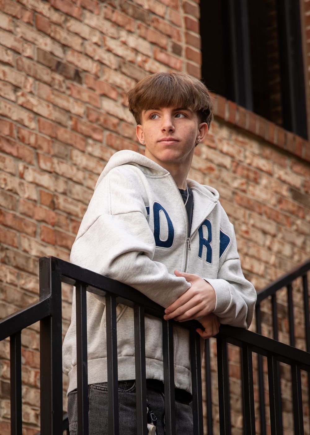 Person in a white hoodie leans against black metal railing with brick wall backdrop in three similar poses.