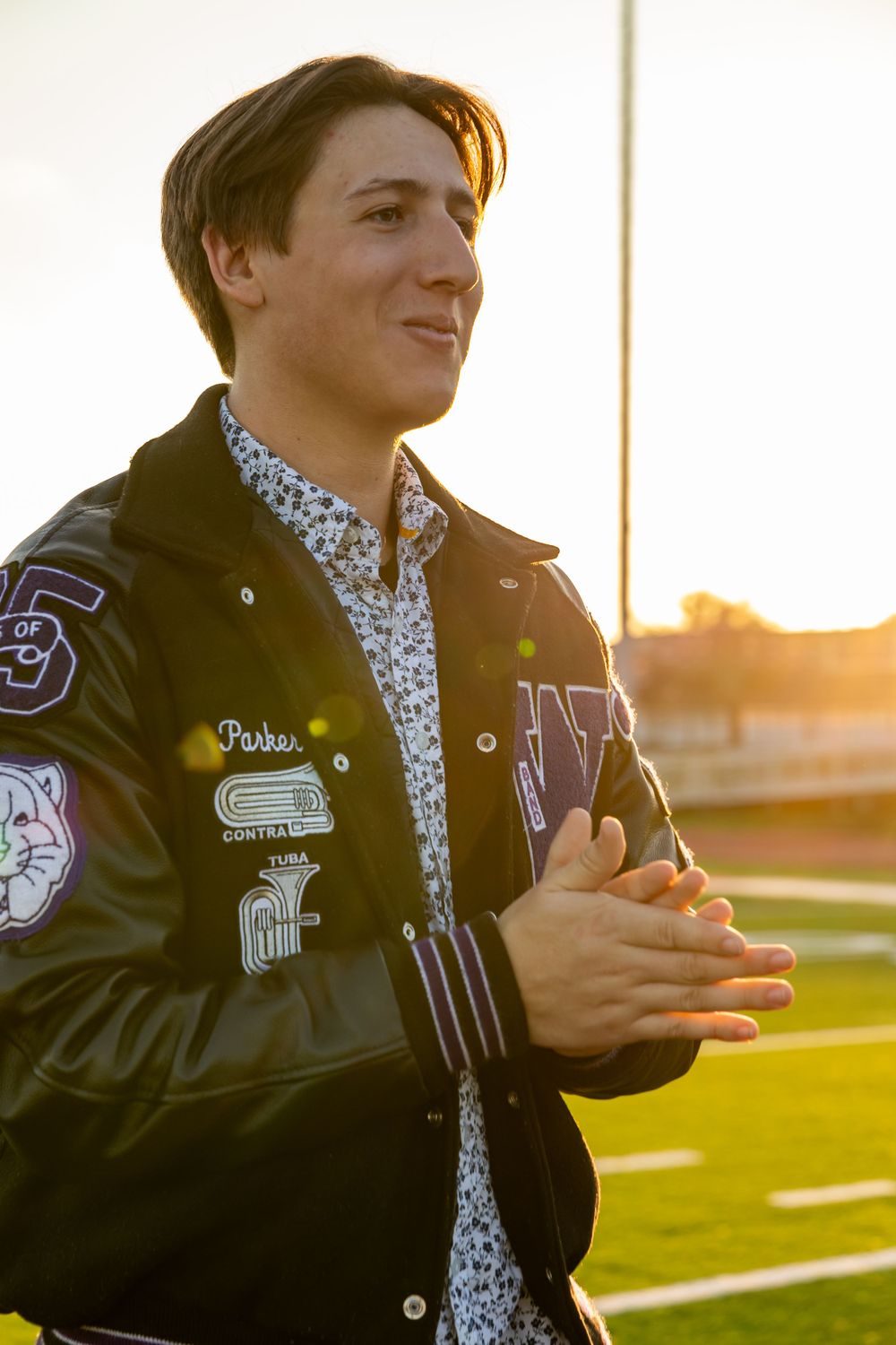 Student in varsity jacket with patches standing on sports field at sunset.
