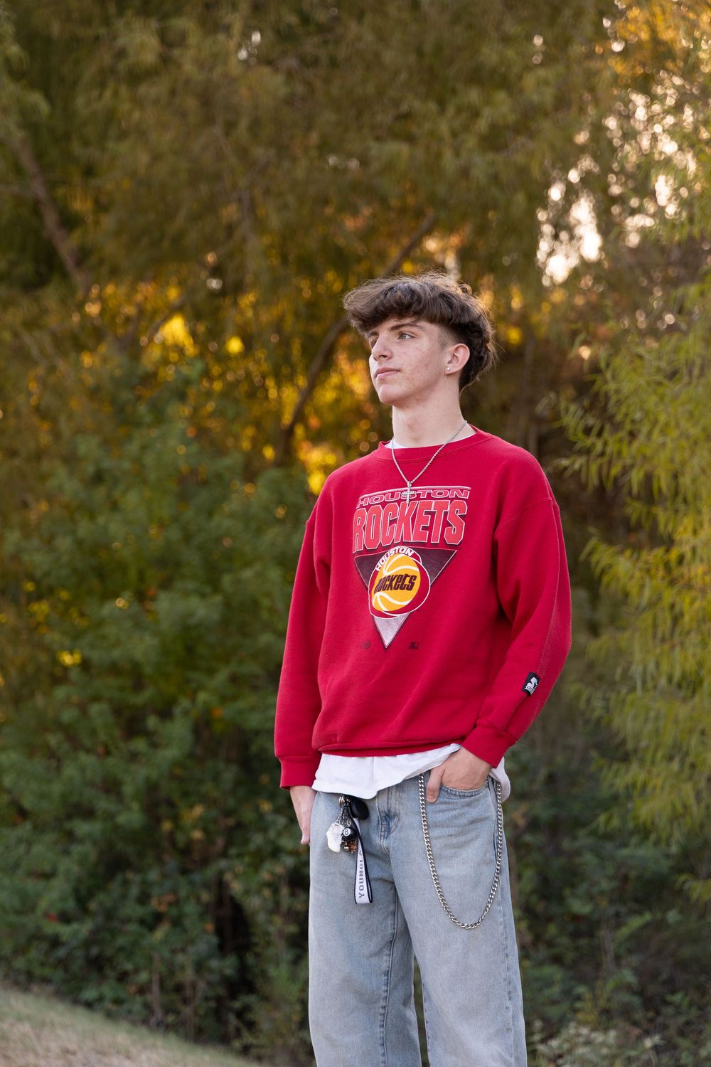 A person wearing a red Houston Rockets sweatshirt and gray sweatpants poses in an outdoor setting with autumn foliage.