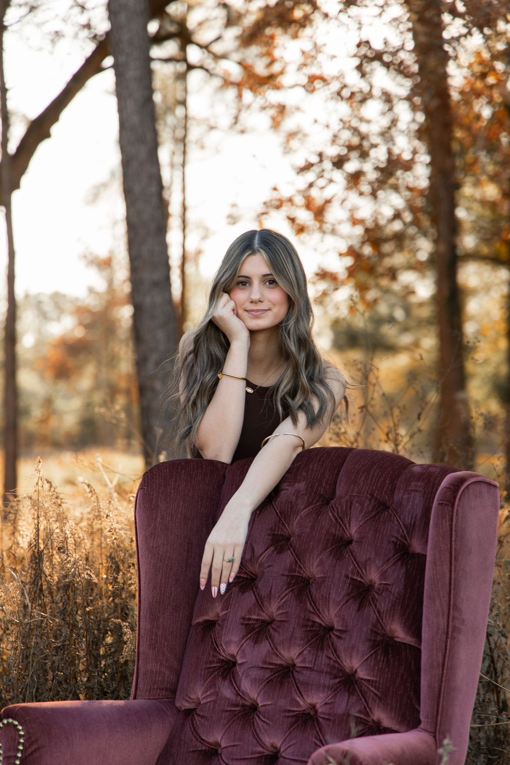 Young woman with wavy hair sits in a tufted burgundy velvet chair outdoors among autumn trees.
