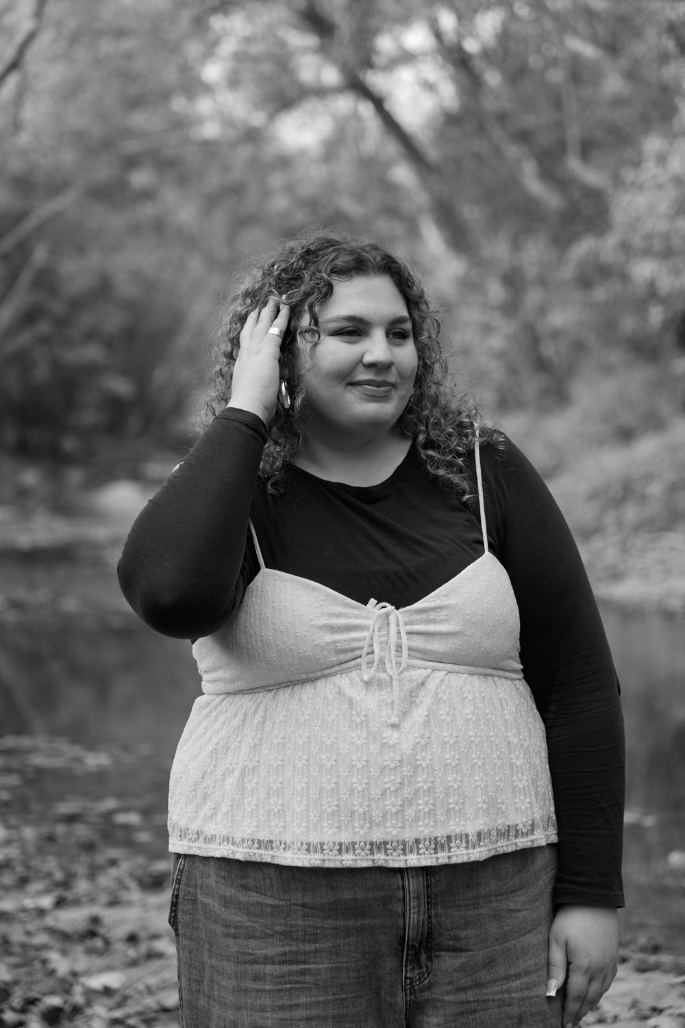A black and white portrait of someone in a black long sleeve top and white camisole standing outdoors among trees.