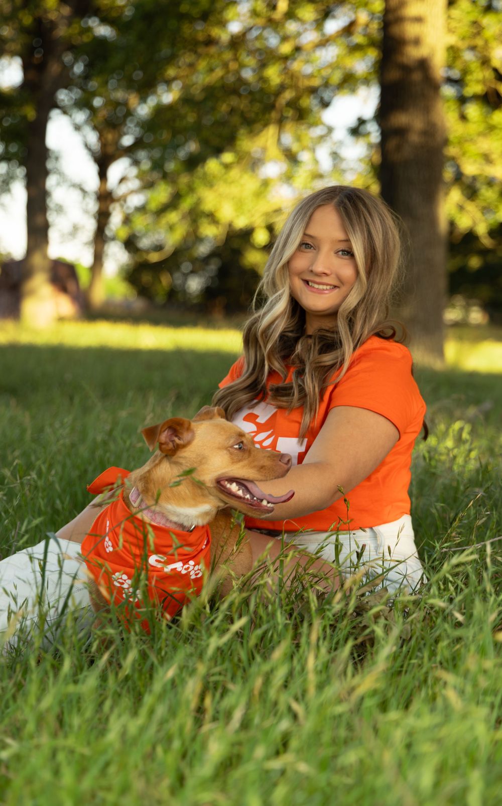 Young woman in orange shirt smiles with golden dog wearing orange bandana, sitting in lush green grass near trees.