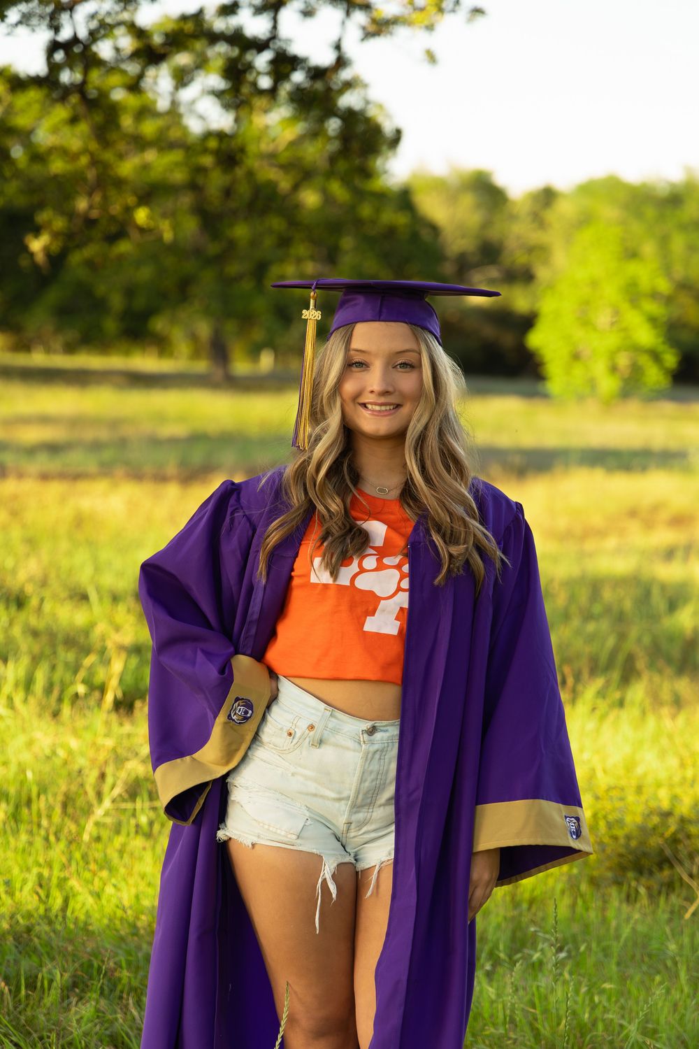 Young woman smiling in purple graduation cap and gown over orange Clemson shirt and denim shorts outdoors.