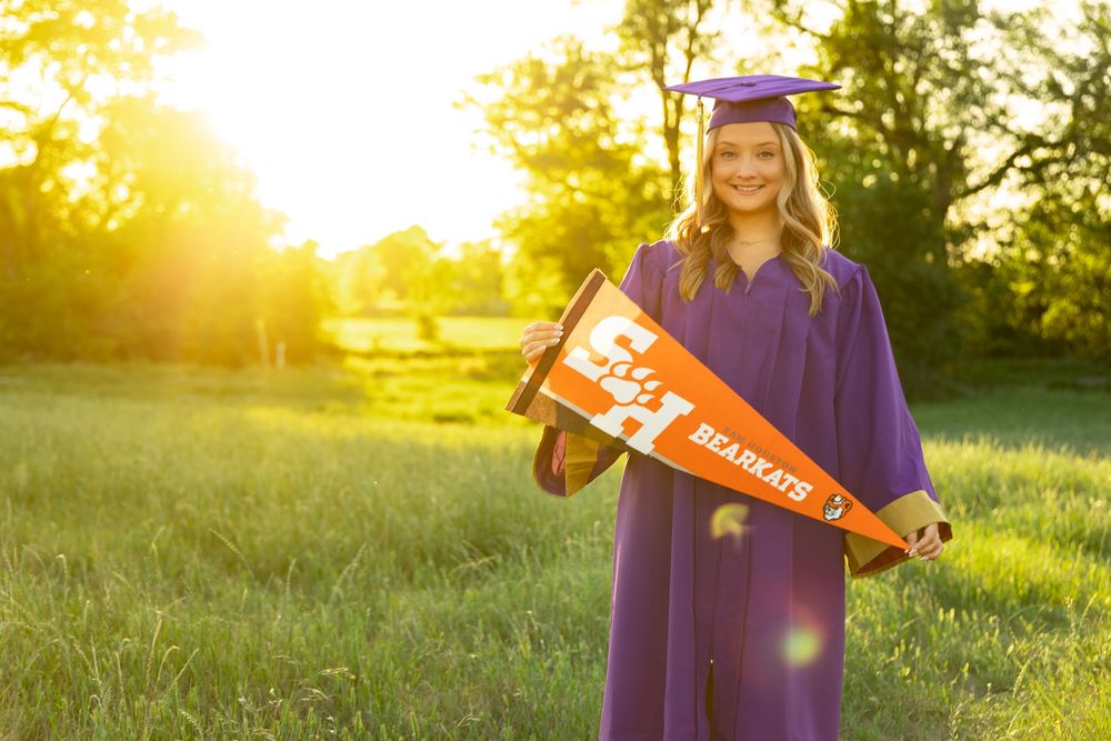 Graduate in purple cap and gown holds Sam Houston State Bearkats orange pennant in sunny green field.