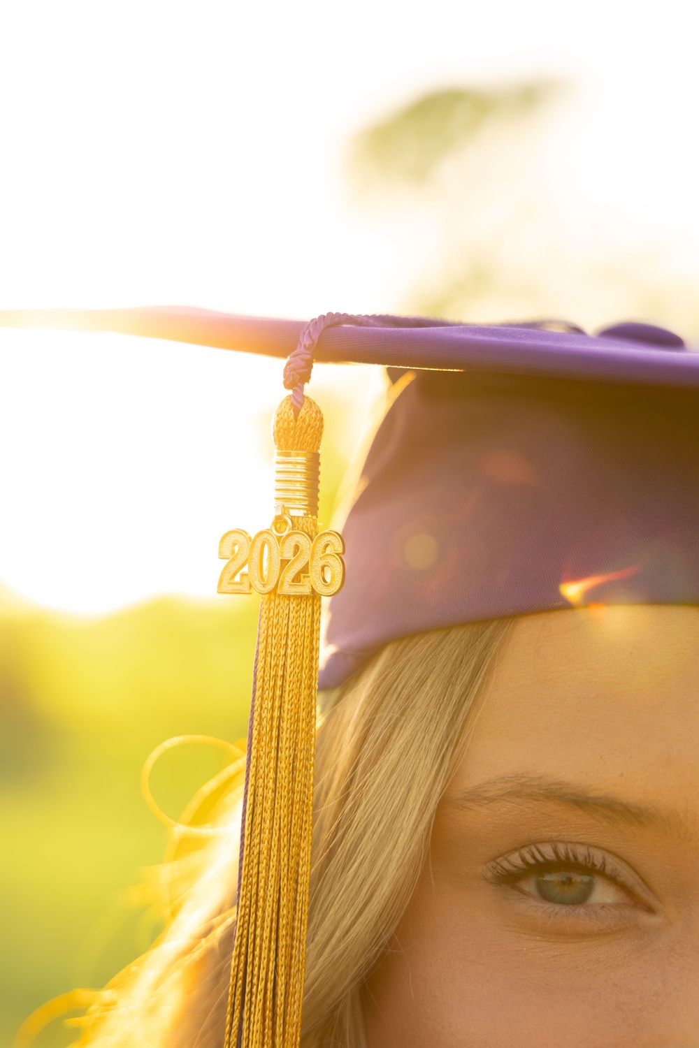 A blonde woman wearing a purple graduation cap with a gold 2026 tassel, photographed outdoors in warm sunlight.