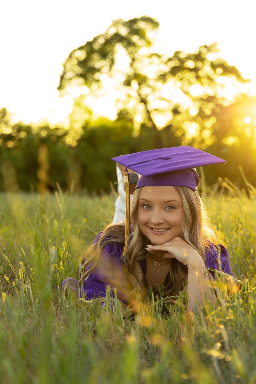 Young woman in purple graduation cap and gown lying in green grass field at golden sunset hour.