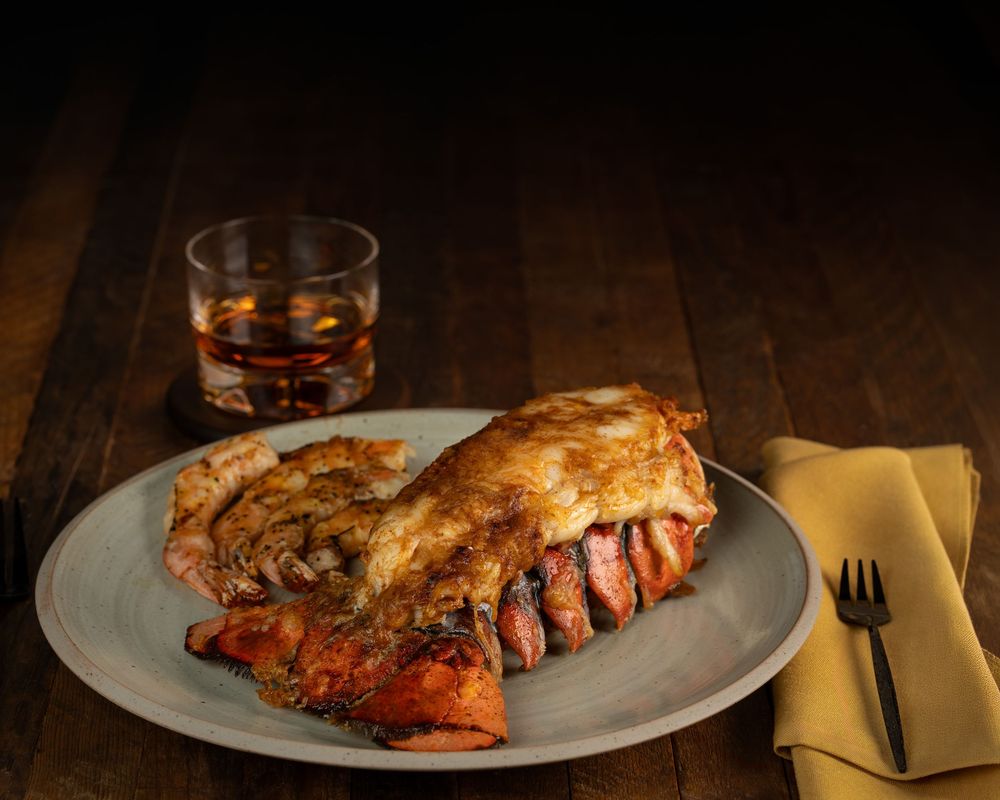 Grilled lobster tail served on a plate with a glass of whiskey and yellow napkin on a dark wooden table.